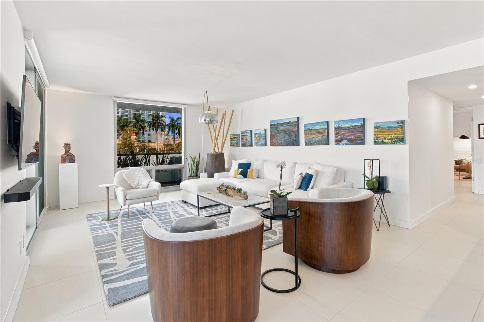 Bright condo living room with a white sectional, curved wood chairs, colorful artwork, and a palm-lined view.