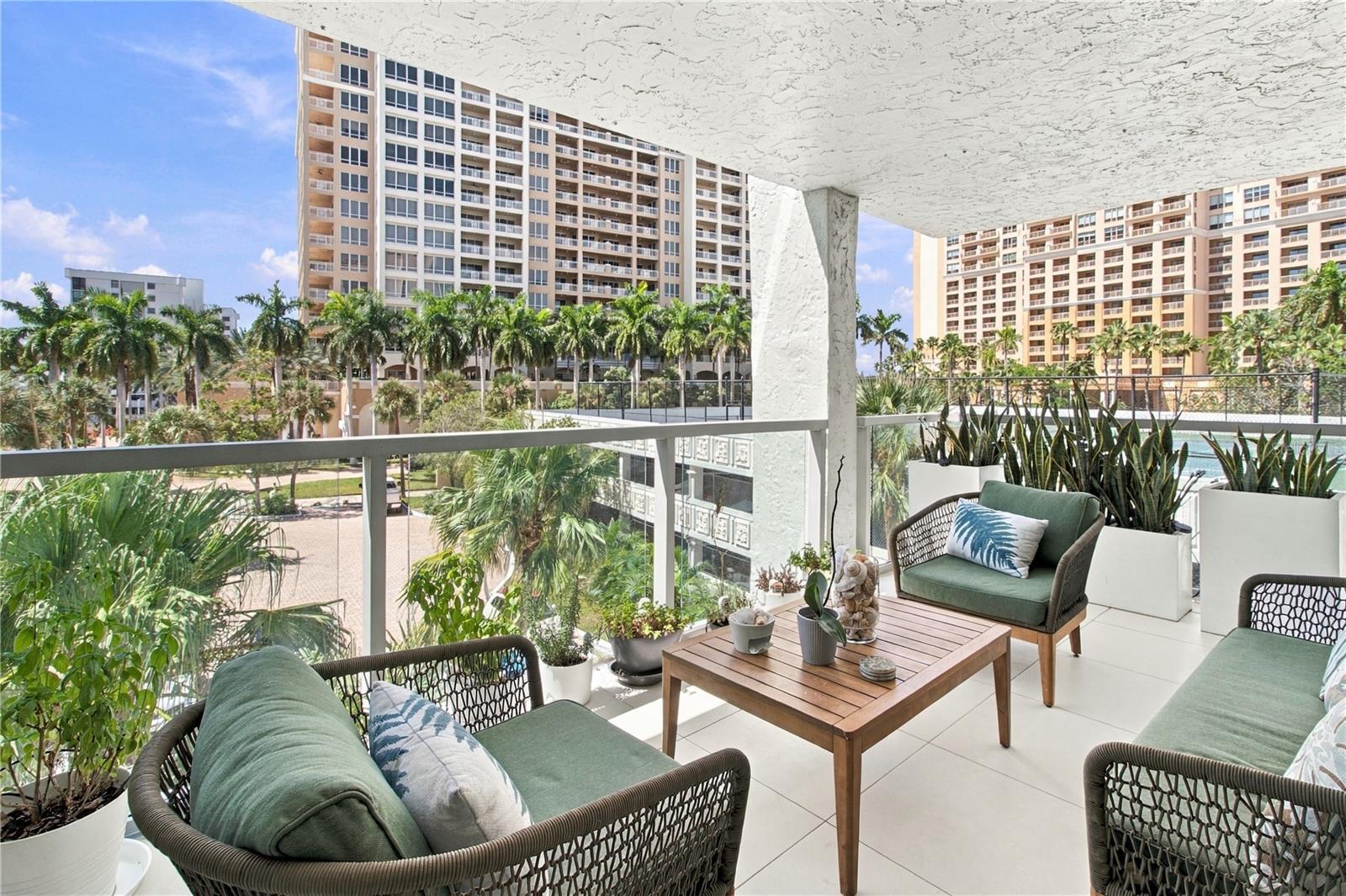 Furnished balcony with wicker seating and potted plants overlooking palm trees and high-rise buildings.
