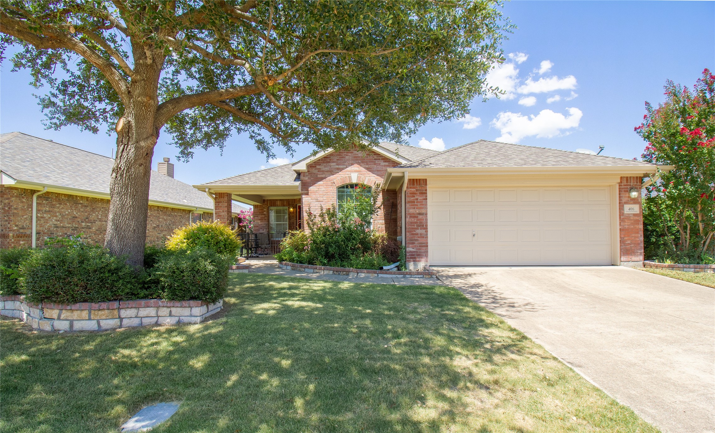 Red-brick home with a two-car garage, shaded by a large tree and surrounded by landscaping.