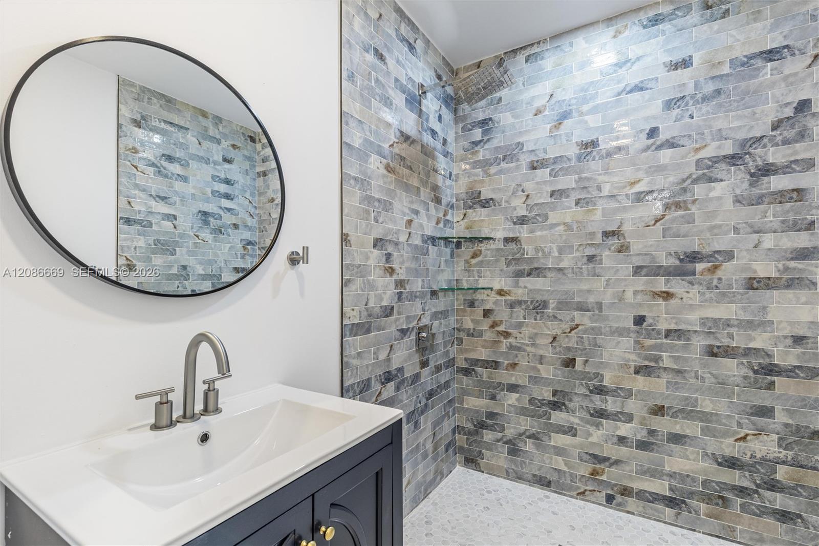 Bathroom vanity and round mirror beside a walk-in shower with gray-blue tile and glass shelves.