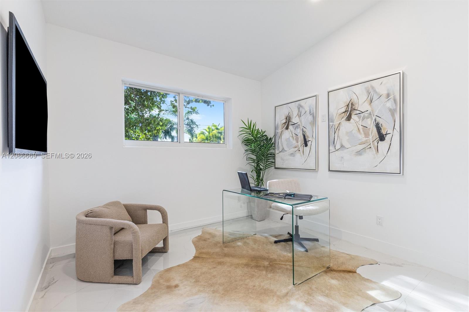 Bright home office with a glass desk, beige armchair, and window overlooking greenery.