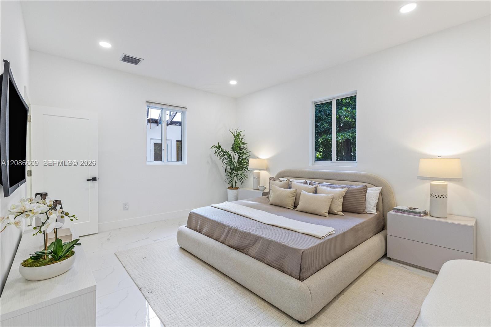 Staged bedroom with a large upholstered bed, neutral bedding, and two windows overlooking greenery.