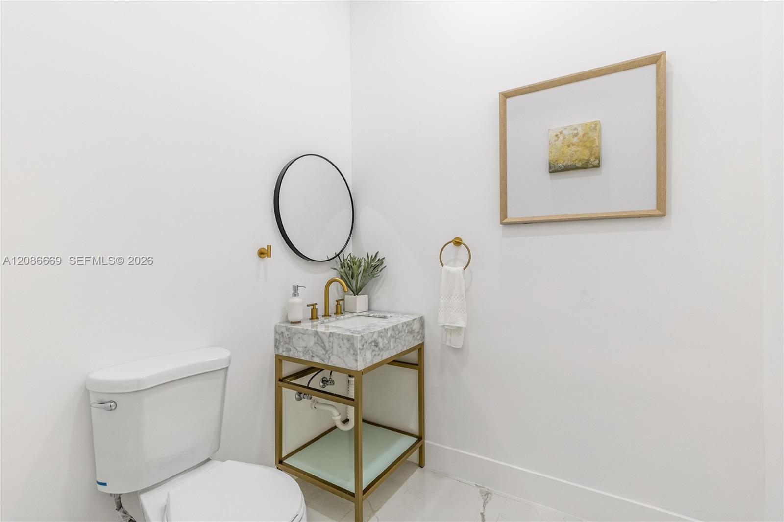 Bright bathroom with marble vanity, gold fixtures, round mirror, and white toilet.