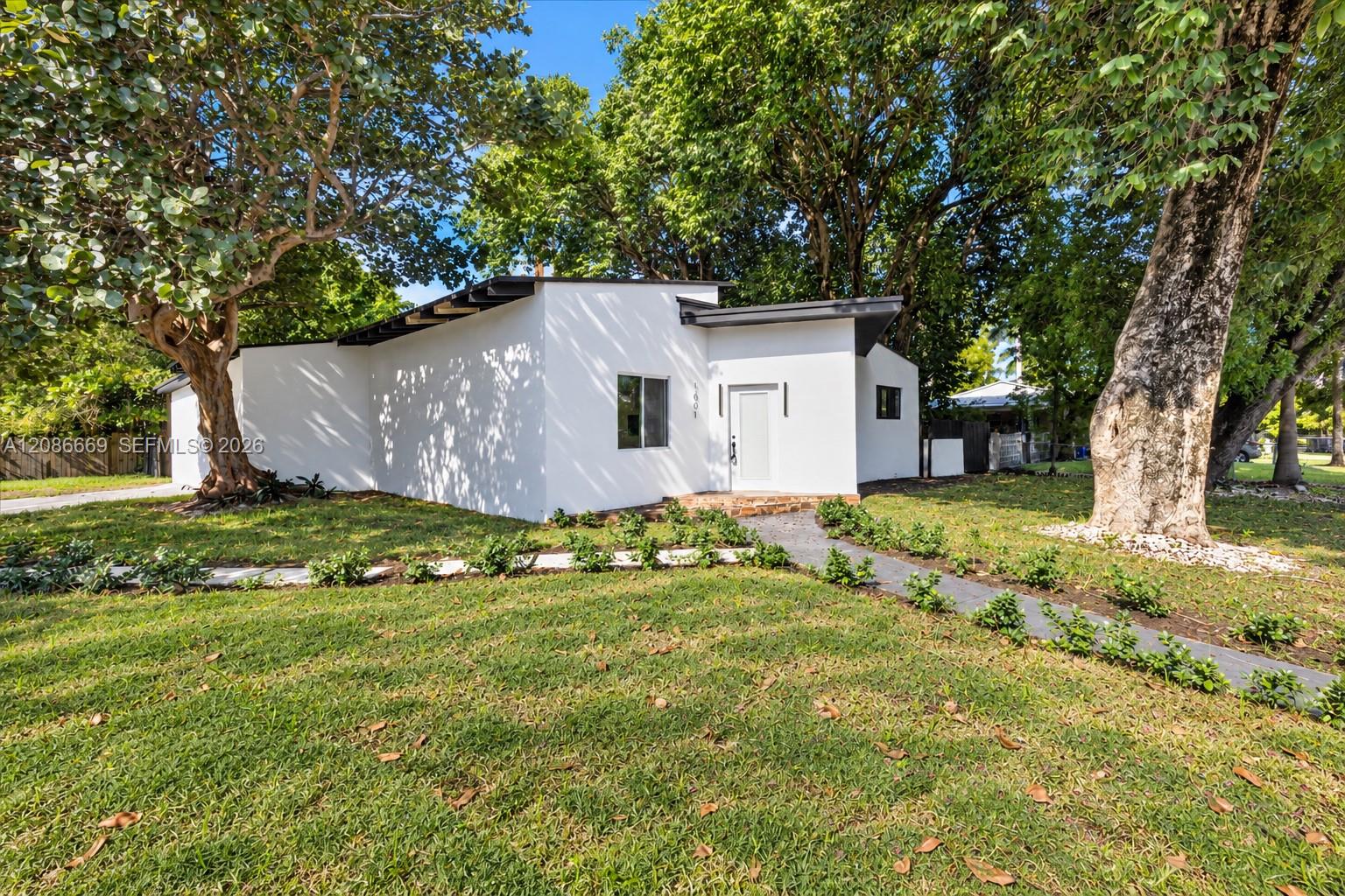 Modern white single-story home framed by mature trees, with a paved path and broad lawn.