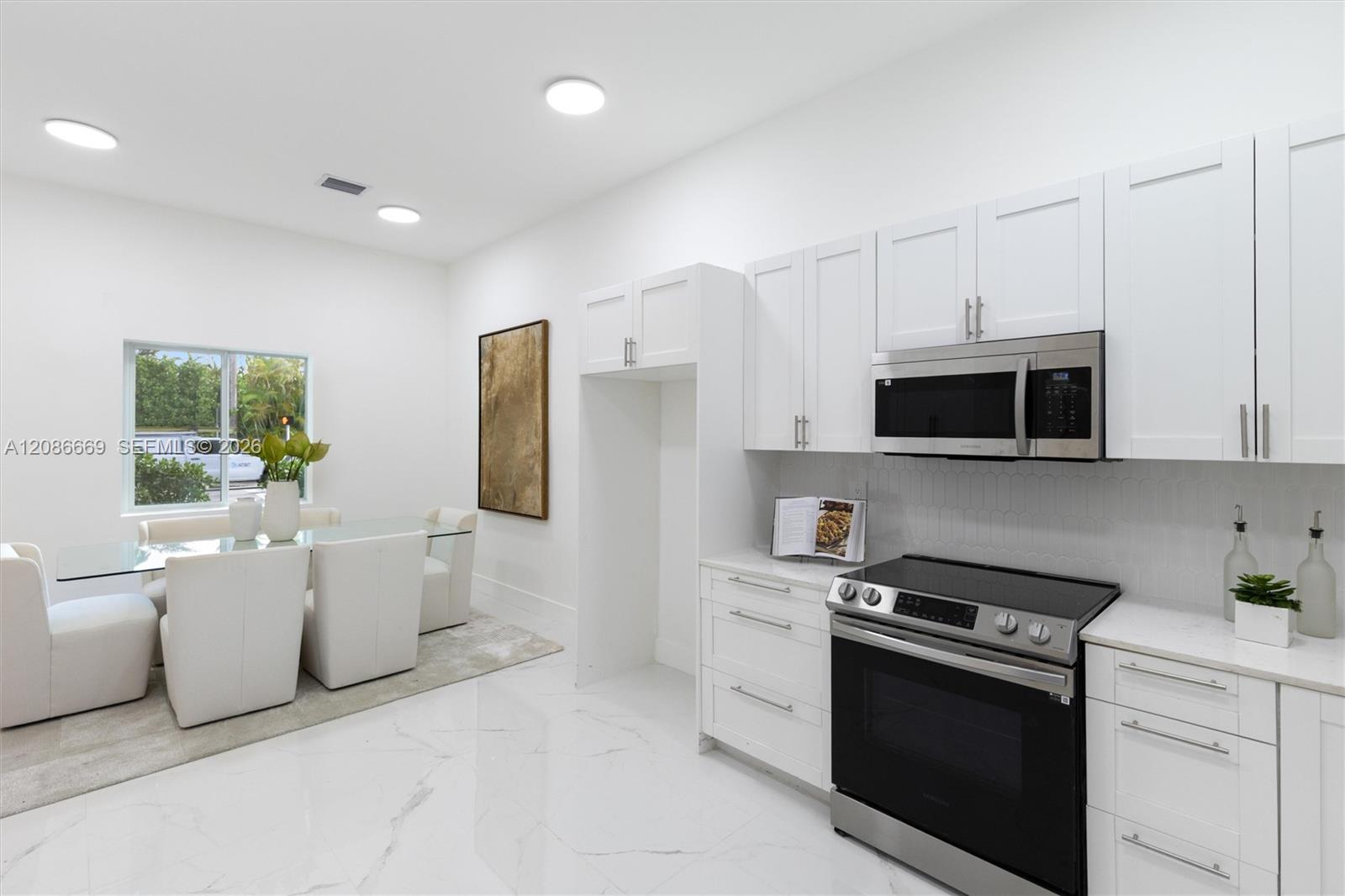 White kitchen with stainless steel appliances beside a dining area with a glass table and white chairs.