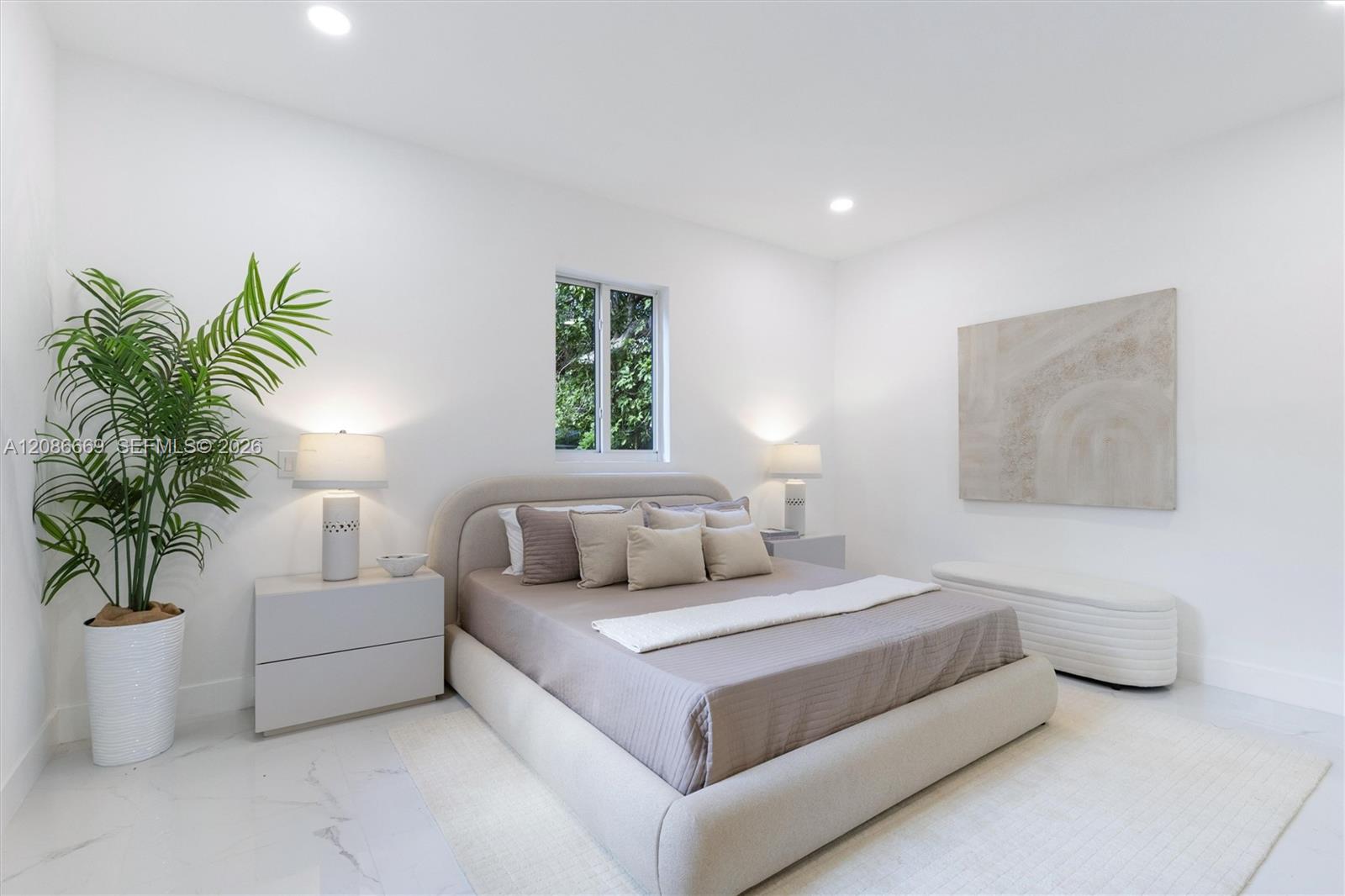 Staged bedroom with a neutral upholstered bed, potted palm, and window overlooking greenery.