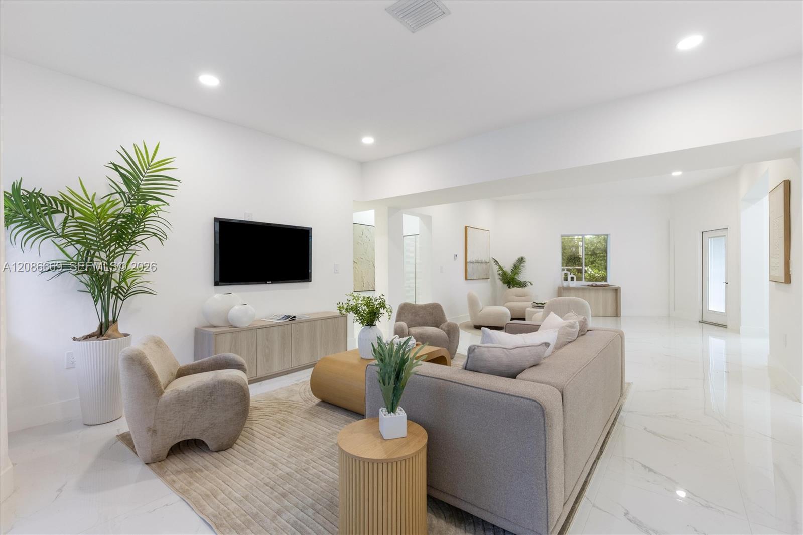 Staged living room with a gray sofa, neutral armchairs, wall-mounted TV, and large potted palm.