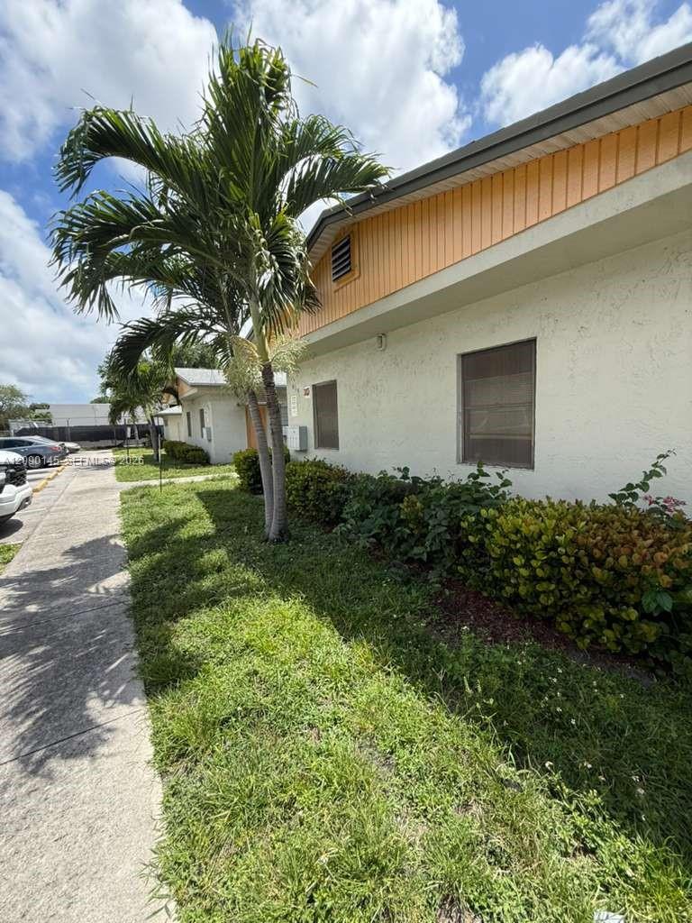 Low-rise property with tan upper siding, palm trees, shrubs, and a sidewalk.