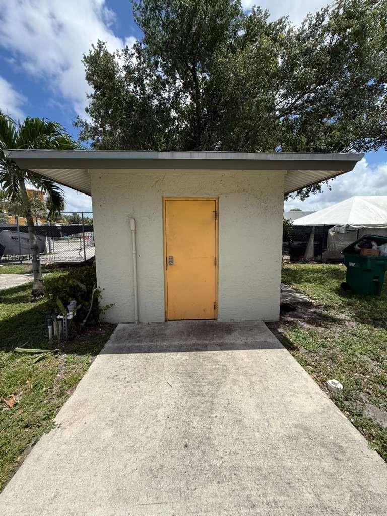 Small white masonry outbuilding with a yellow door and concrete walkway.