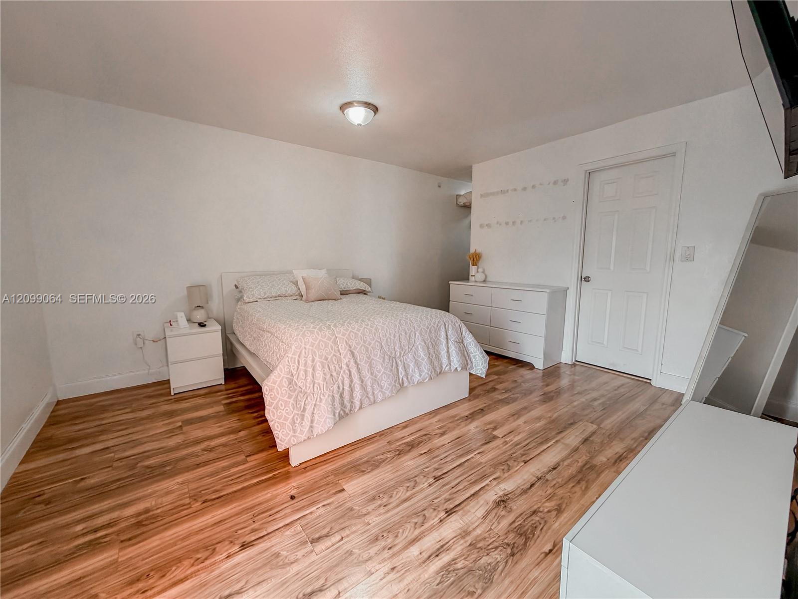 Bedroom with a white bed, pale patterned bedding, white dresser, and wood-look flooring.