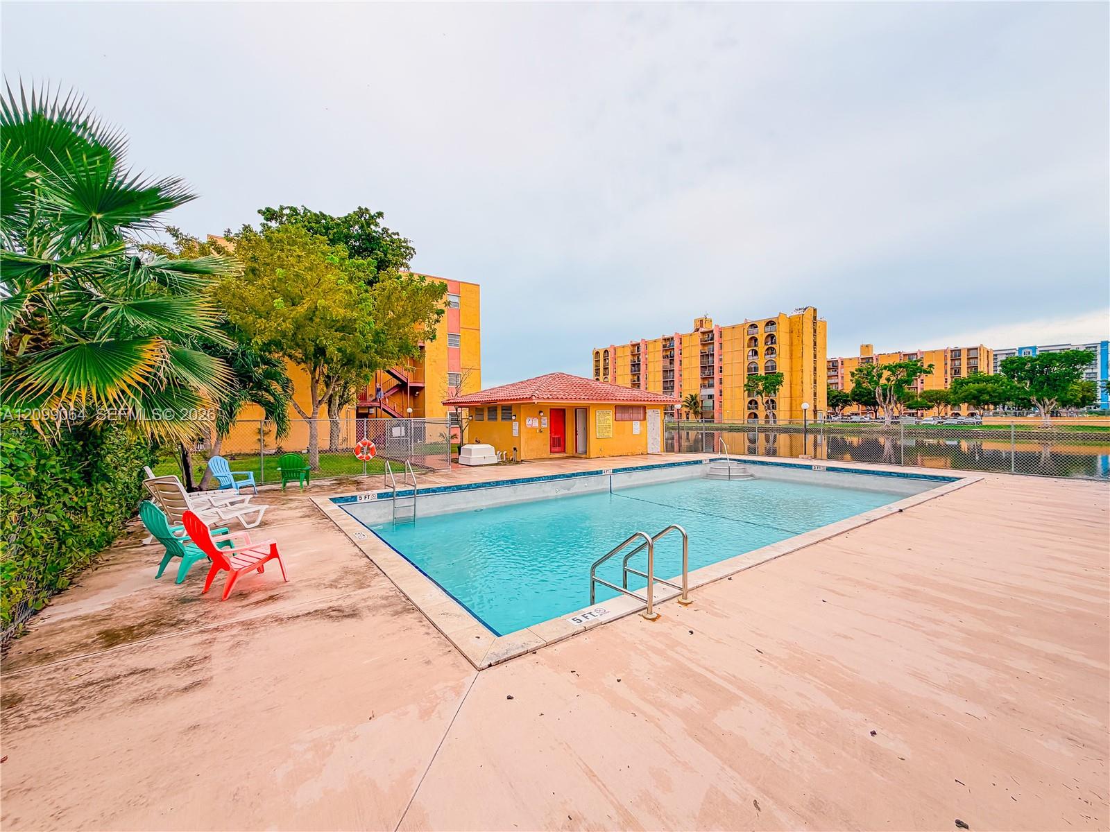 Apartment complex pool with lounge chairs, a low pool building, and multistory buildings behind it.