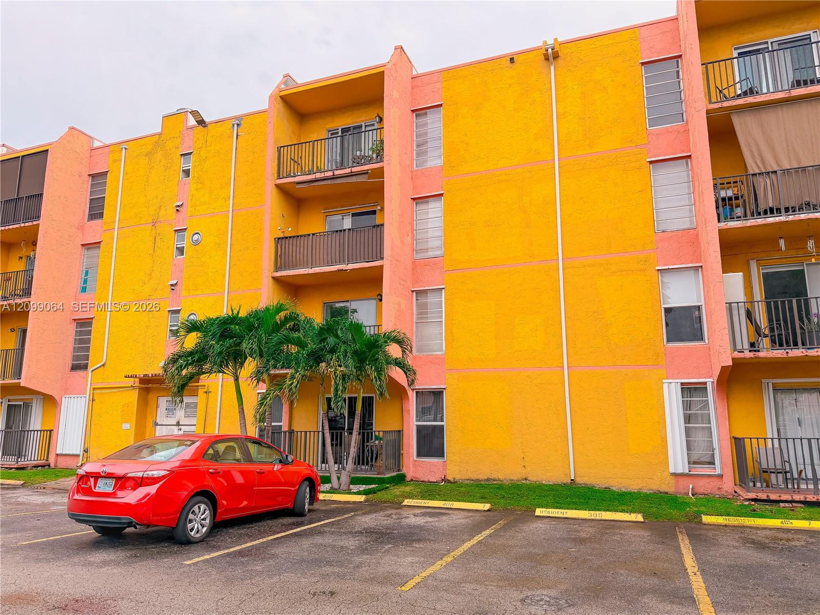 Yellow apartment building with coral trim, recessed balconies, palm trees, and a red car parked outside.