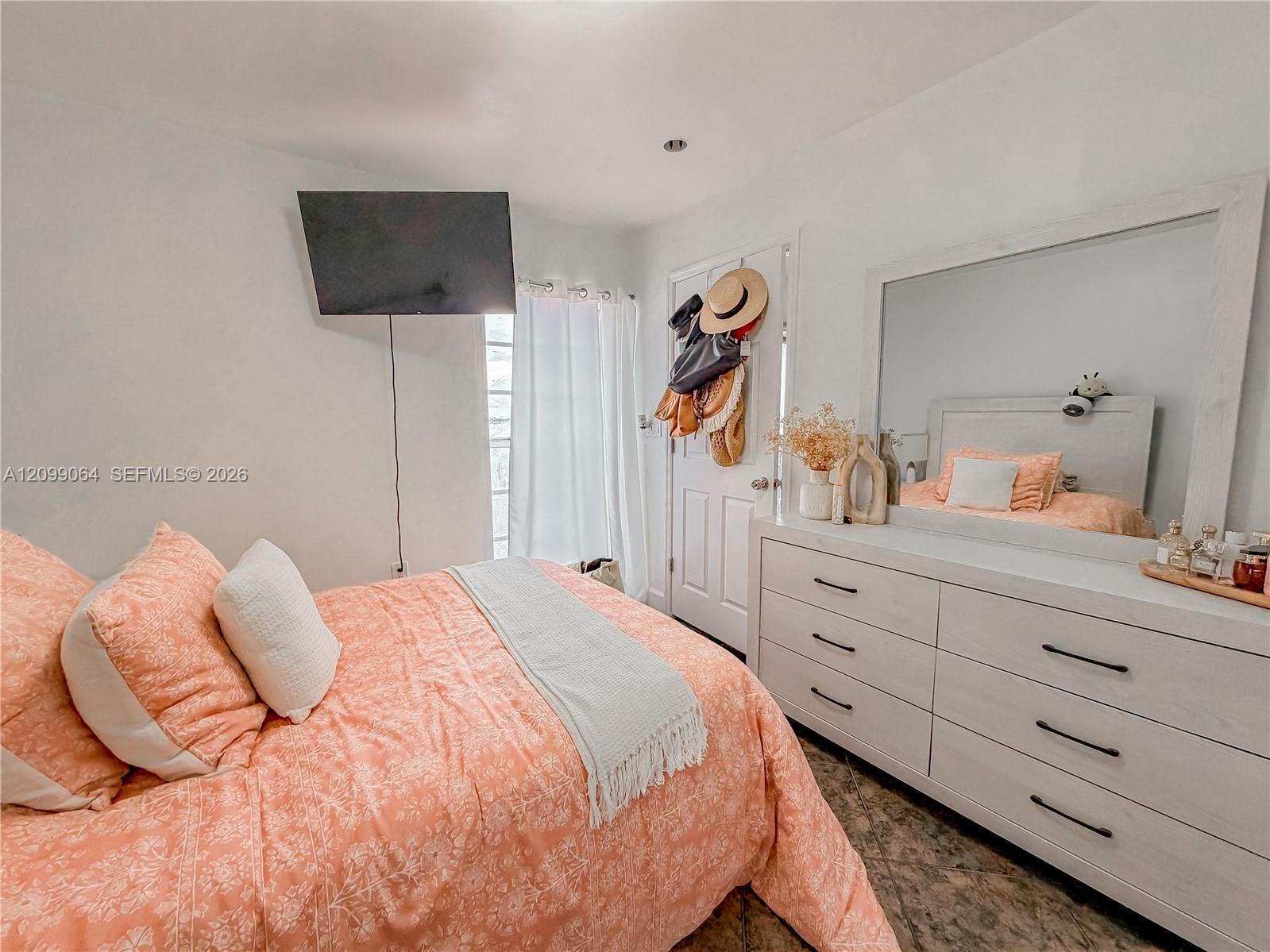 Bedroom with a peach patterned bedspread, white dresser and mirror, and wall-mounted television.