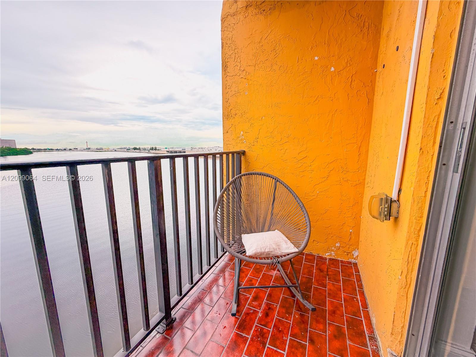 Balcony overlooking water, with red tile flooring, an orange stucco wall, and a woven chair.
