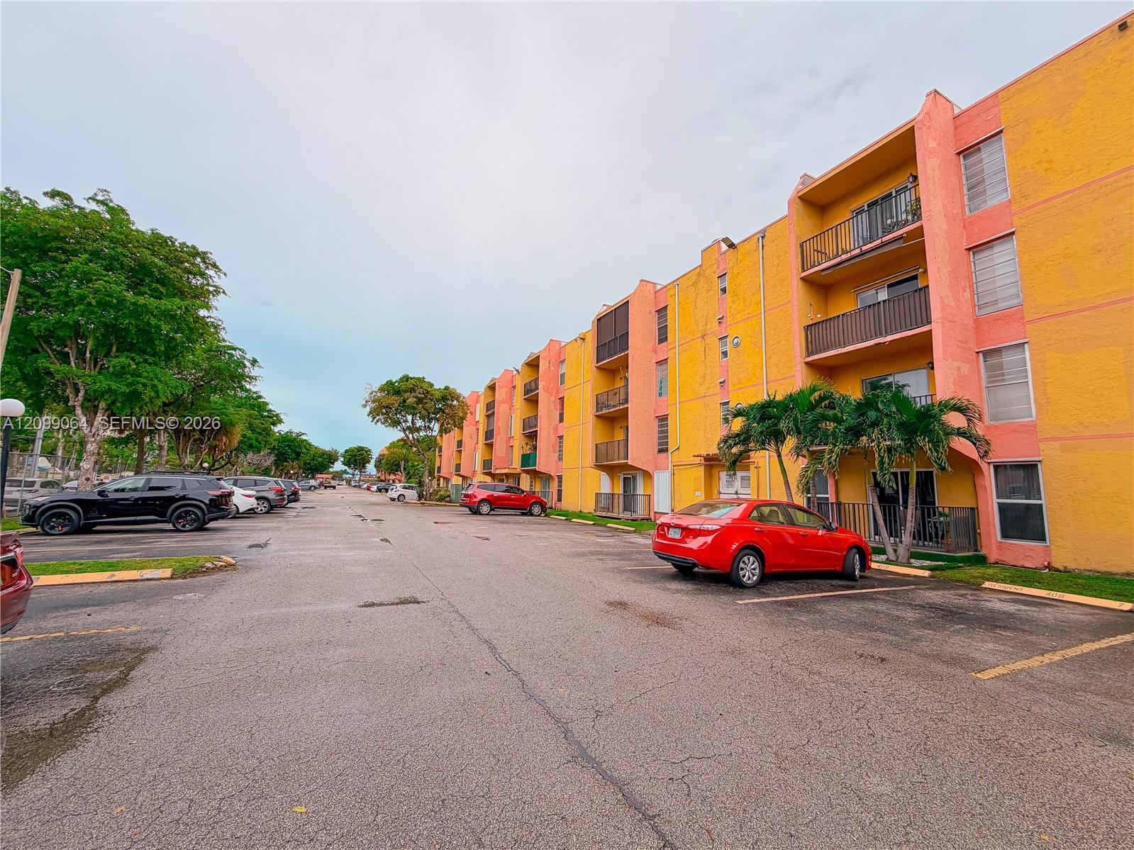 Apartment complex with yellow and coral walls, balconies, palm trees, and a parking lot.