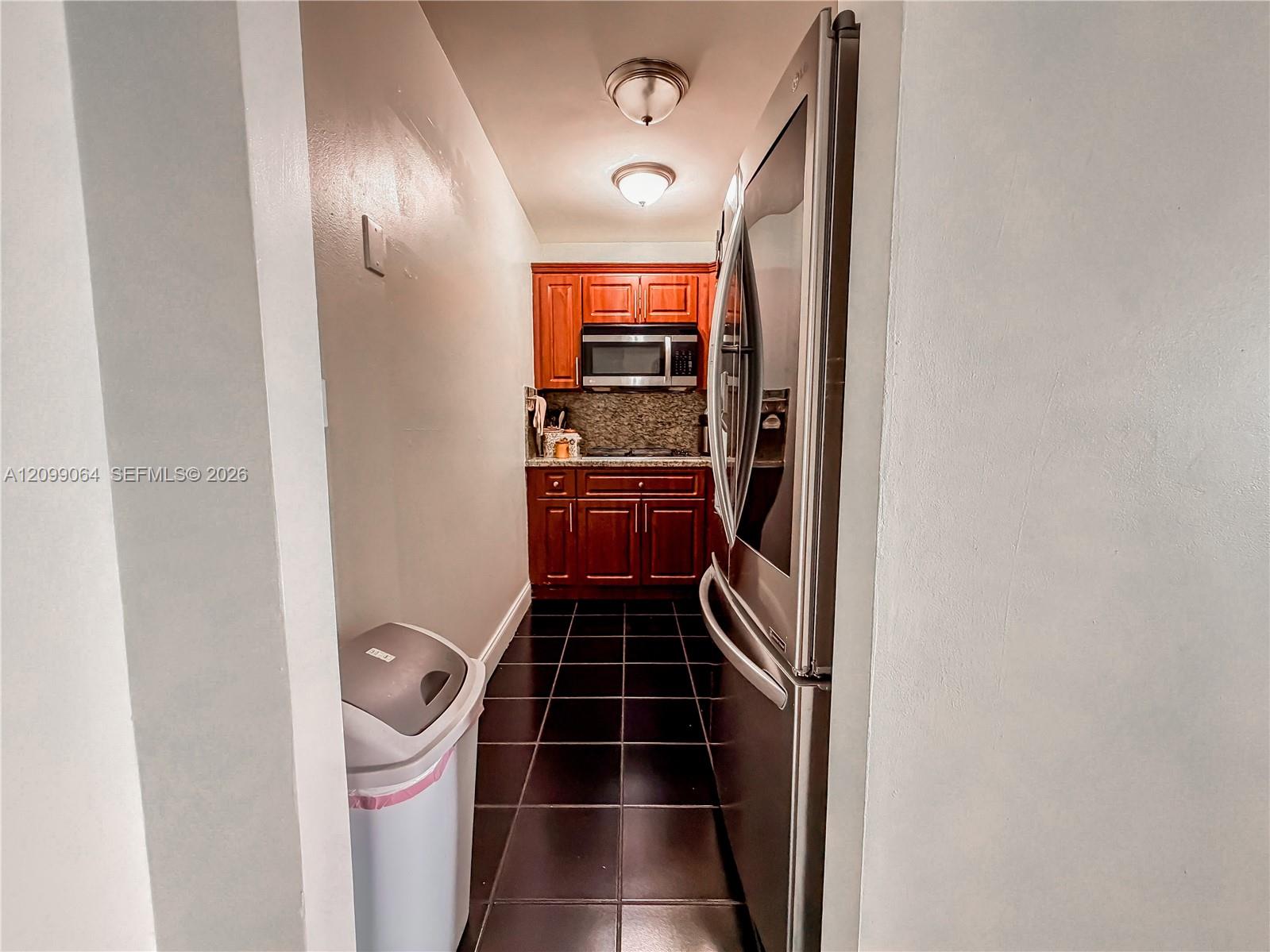 Narrow galley kitchen with wood cabinets, dark tile flooring, and stainless steel appliances.