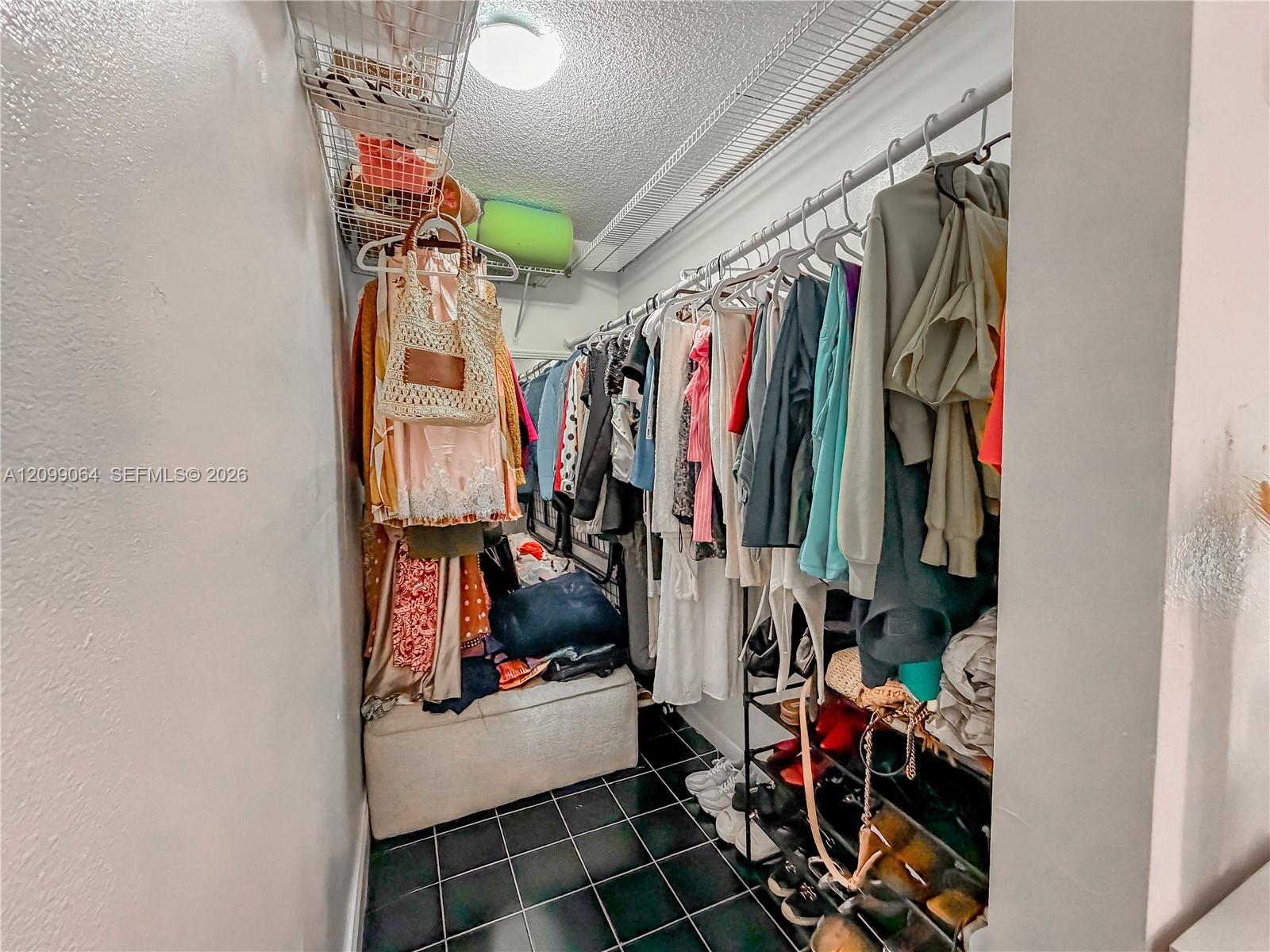 Walk-in closet with clothes on both sides, wire shelving, shoes, and a storage ottoman.