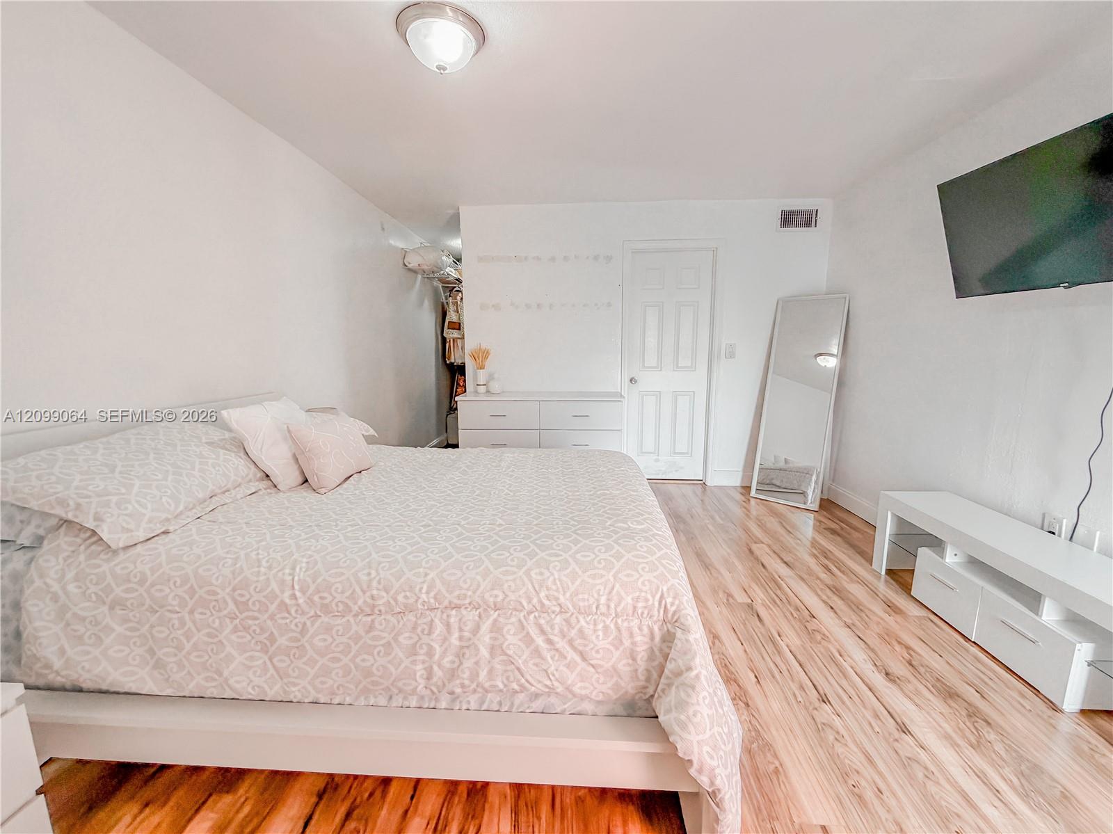 Bedroom with a large patterned bed, wood-look flooring, wall-mounted television, and leaning full-length mirror.