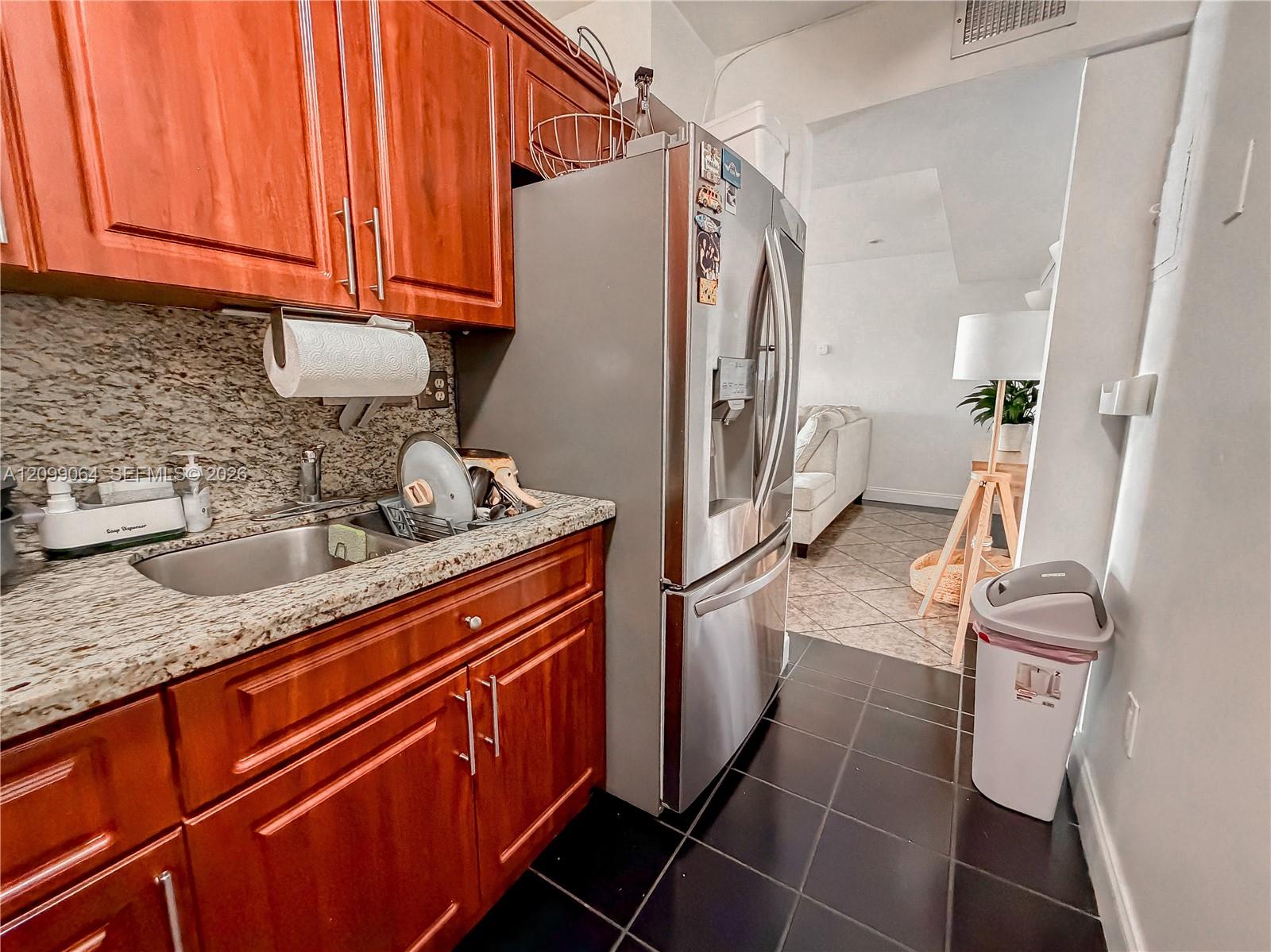Kitchen with reddish wood cabinets, granite counters, and a stainless-steel refrigerator opening to the living area.