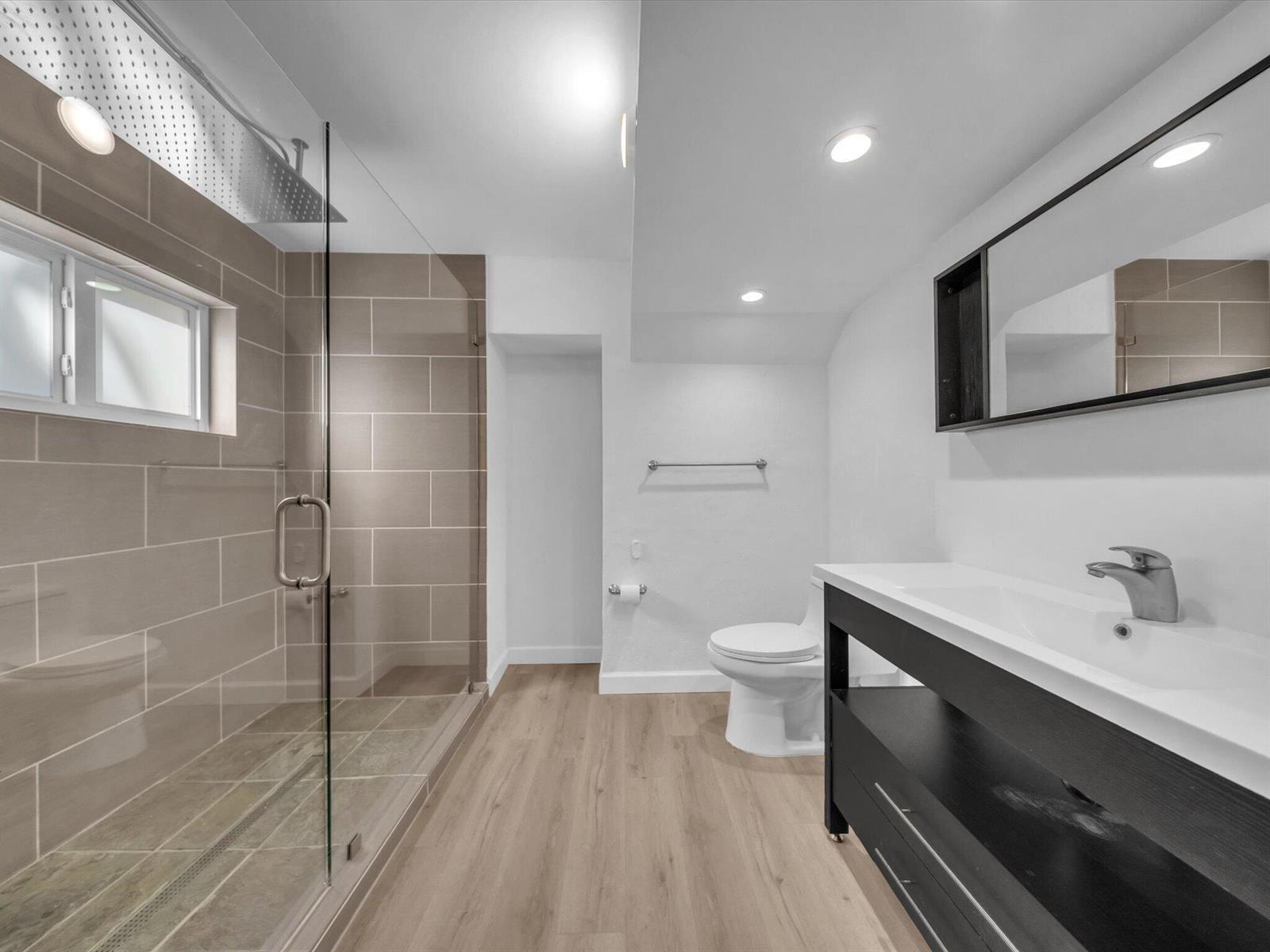 Modern bathroom with a glass walk-in shower, dark vanity, mirrored cabinet, and light wood-look flooring.