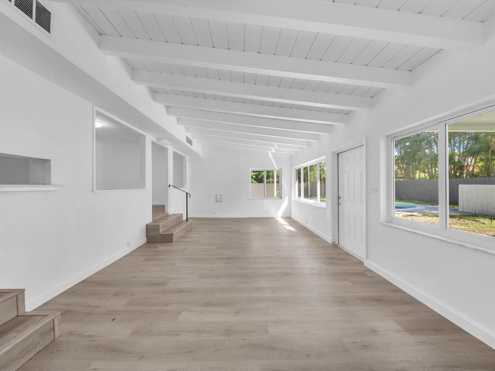 Vacant living area with exposed white ceiling beams, pale floors, and large windows overlooking the yard.