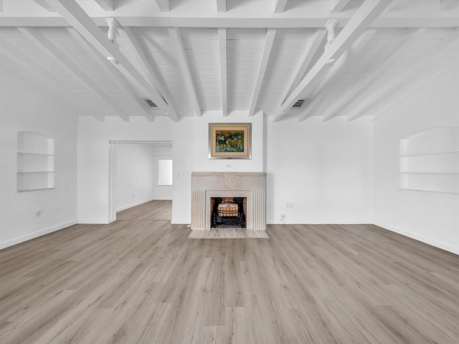 Vacant living room with exposed ceiling beams, built-in shelves, and a central fireplace.