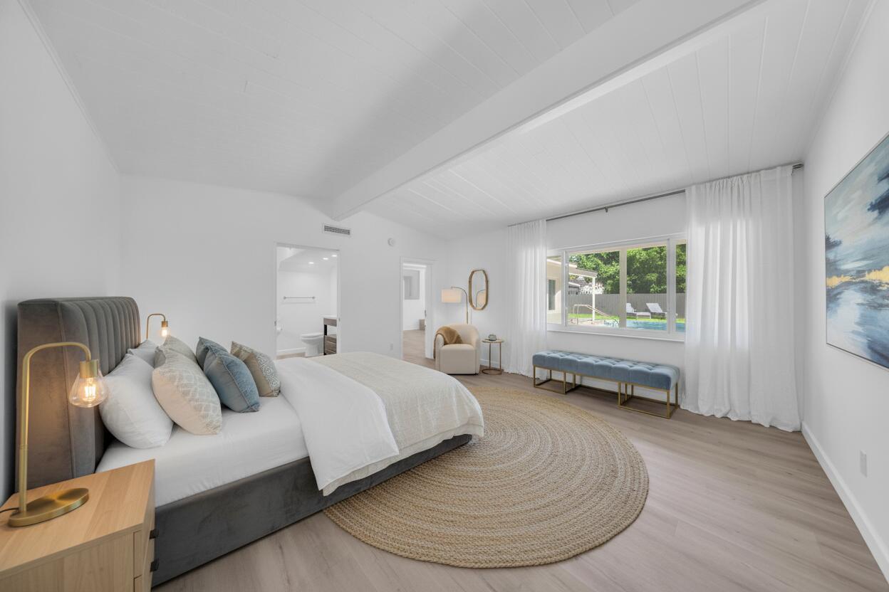 Bedroom with a gray upholstered bed, vaulted white ceiling, and window overlooking a pool.