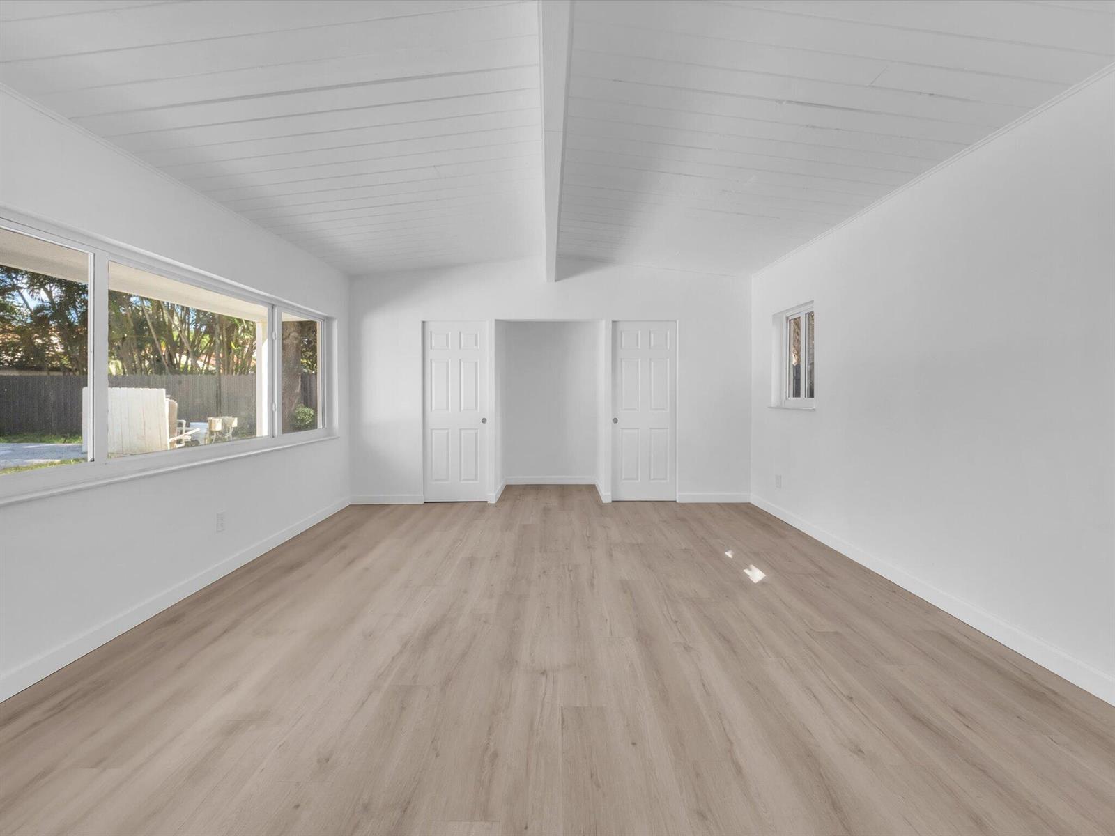 Empty room with a vaulted white ceiling, light wood flooring, and large windows.