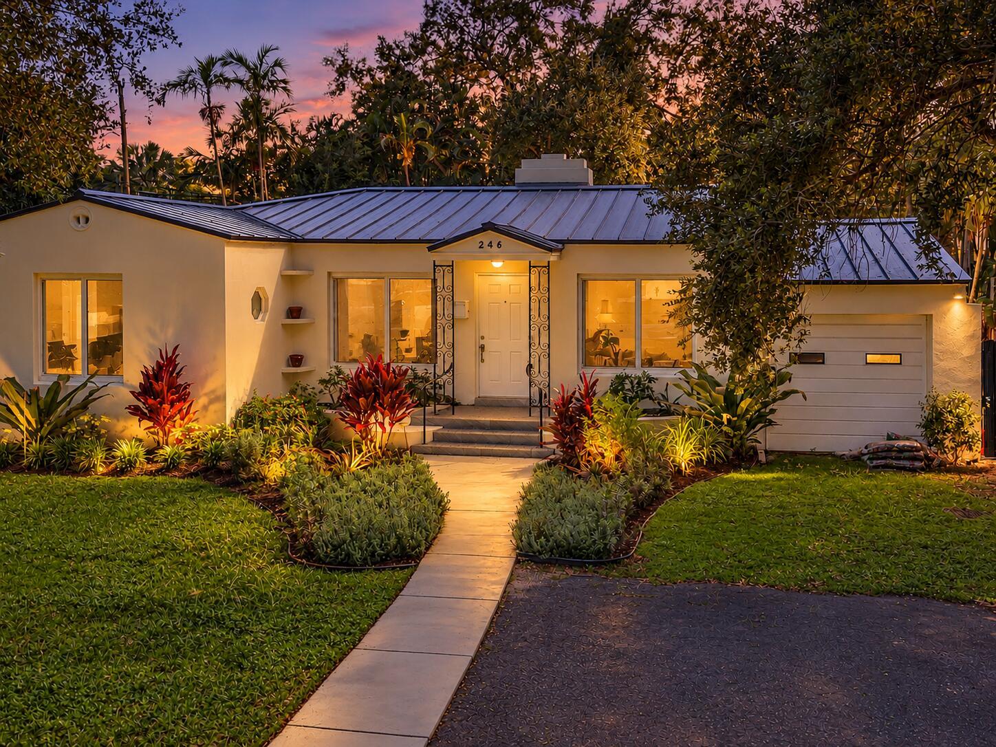 Single-story home with a metal roof, tropical landscaping, and a warmly lit entry at dusk.