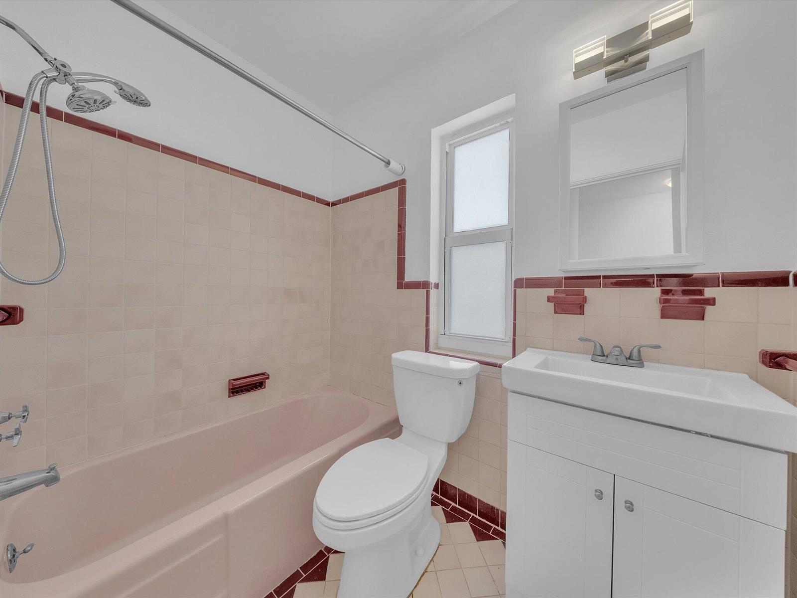 Bathroom with a pink bathtub, burgundy tile trim, white toilet, and vanity.
