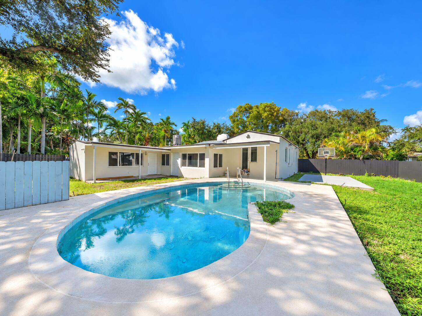 White single-story home behind a curved backyard pool, surrounded by tropical palms and a broad stone patio.