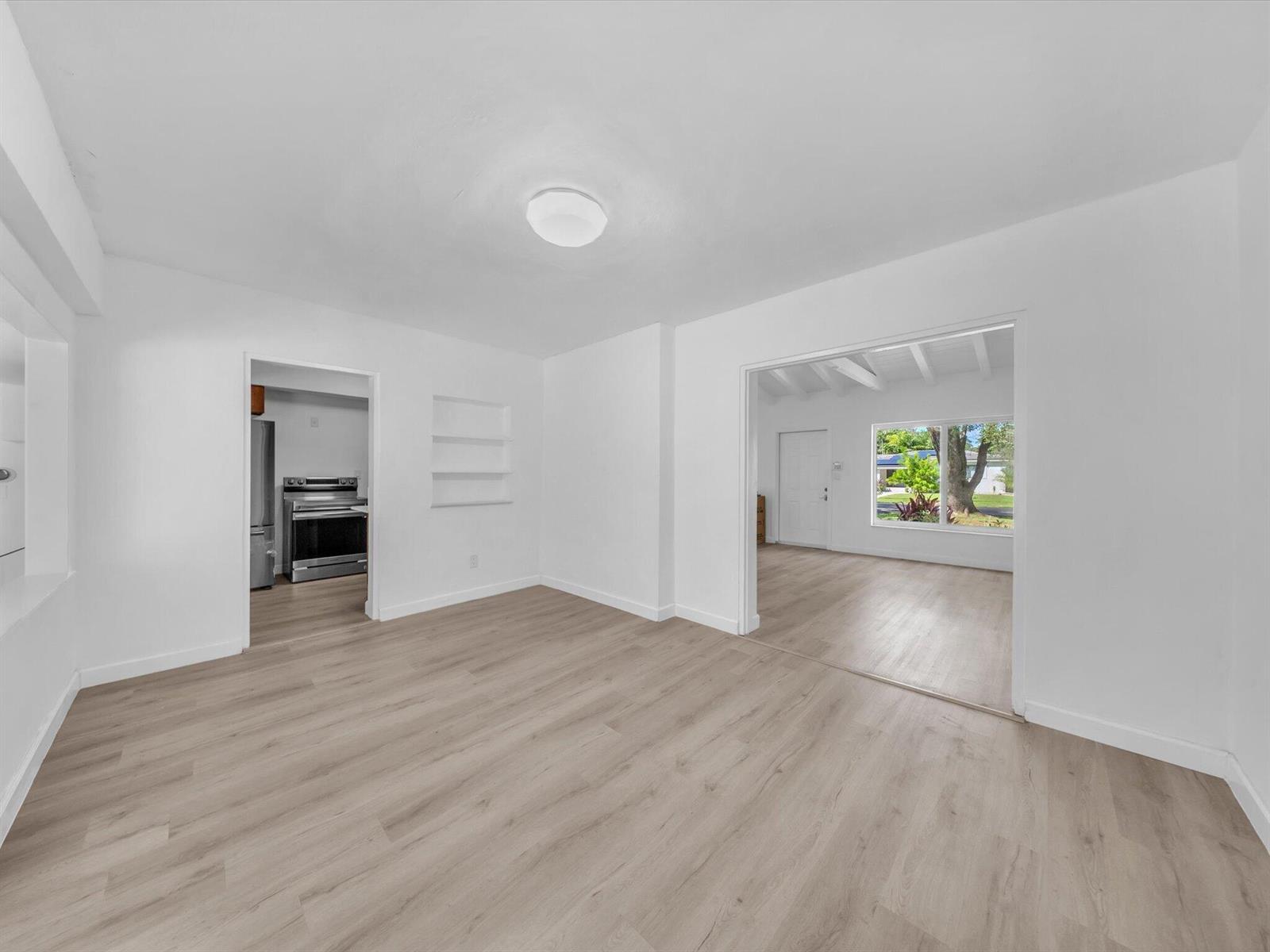 Vacant main room with light wood-look floors, opening to a kitchen and a bright room with a large window.