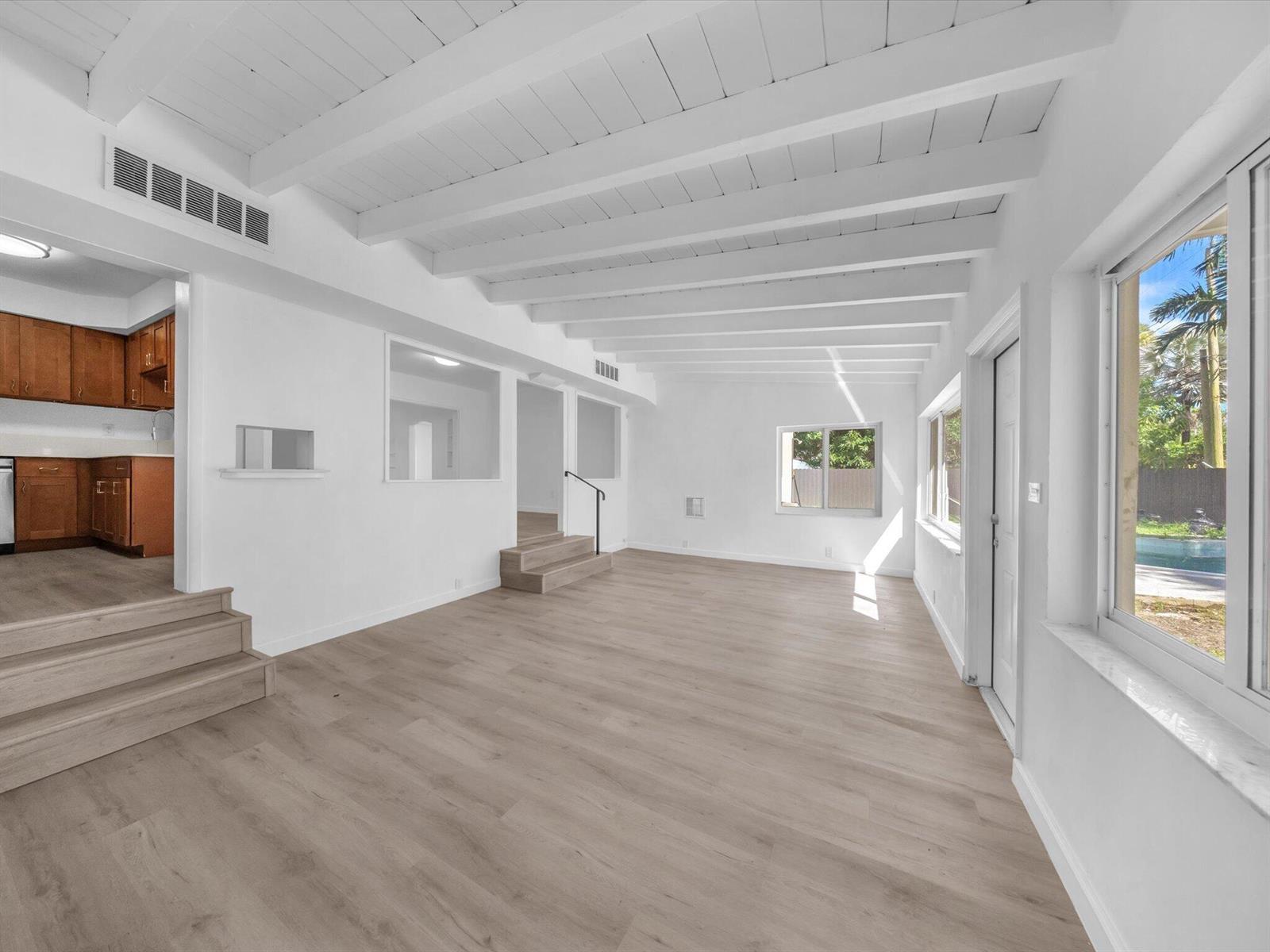 Vacant living room with a vaulted beam ceiling, pale wood floors, and windows overlooking a pool.