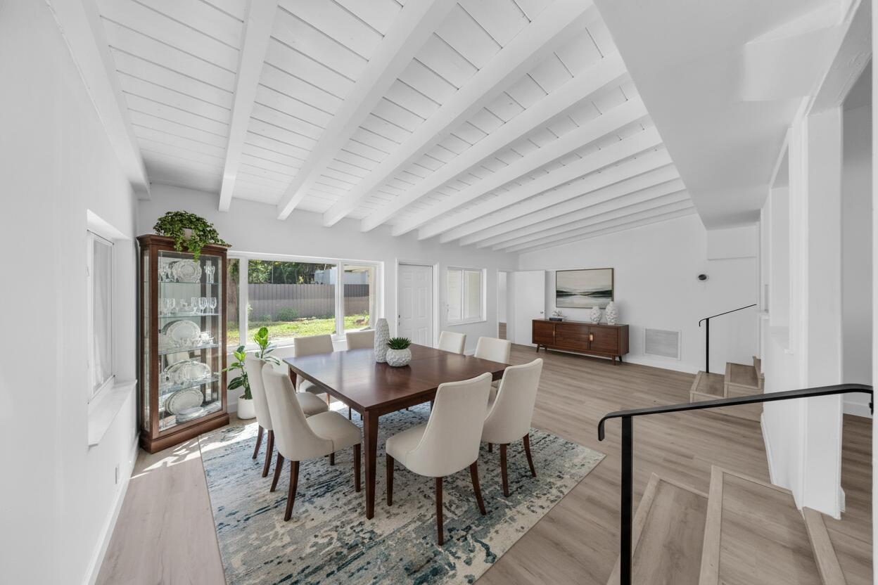 Dining room with exposed white ceiling beams, a dark wood table, and windows overlooking the yard.
