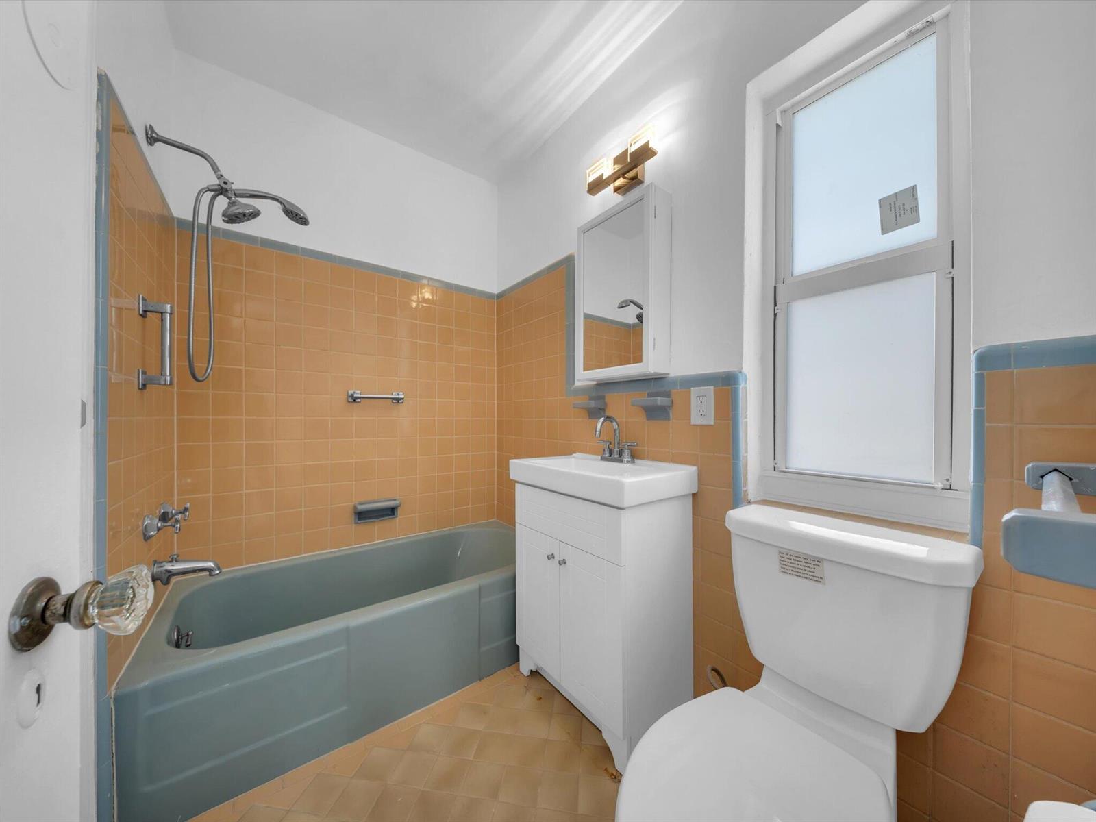 Bathroom with a blue bathtub, orange tile walls, and a frosted window beside the toilet.