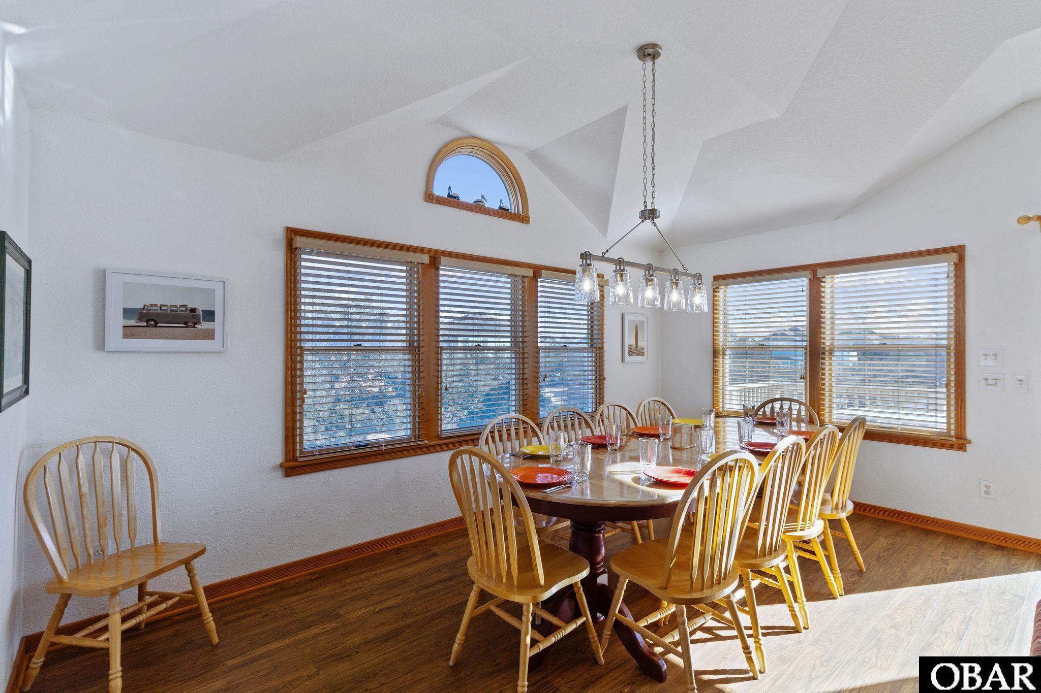 Dining room with a set table, wooden chairs, large windows, and a vaulted ceiling.