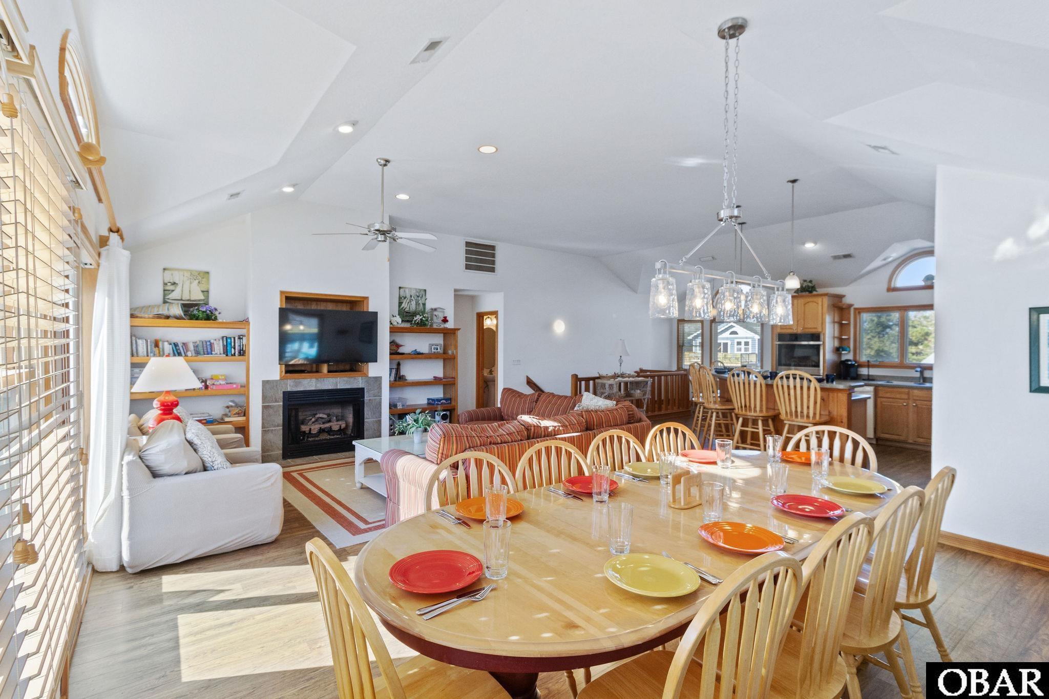 Open-plan living and dining area with a set table, vaulted ceiling, fireplace, and kitchen beyond.