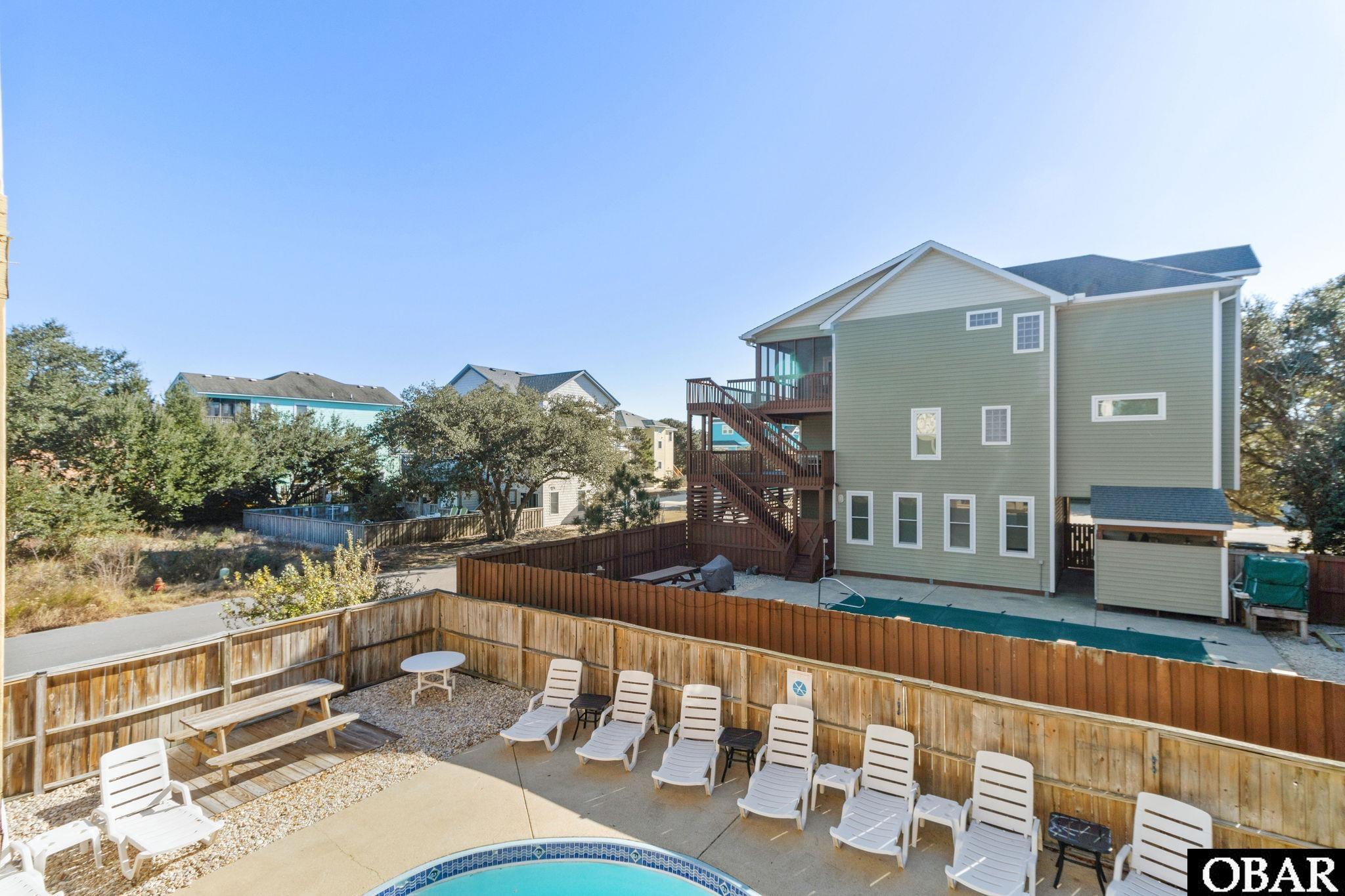 Green multi-story house with wooden decks overlooking a fenced pool and outdoor seating area.