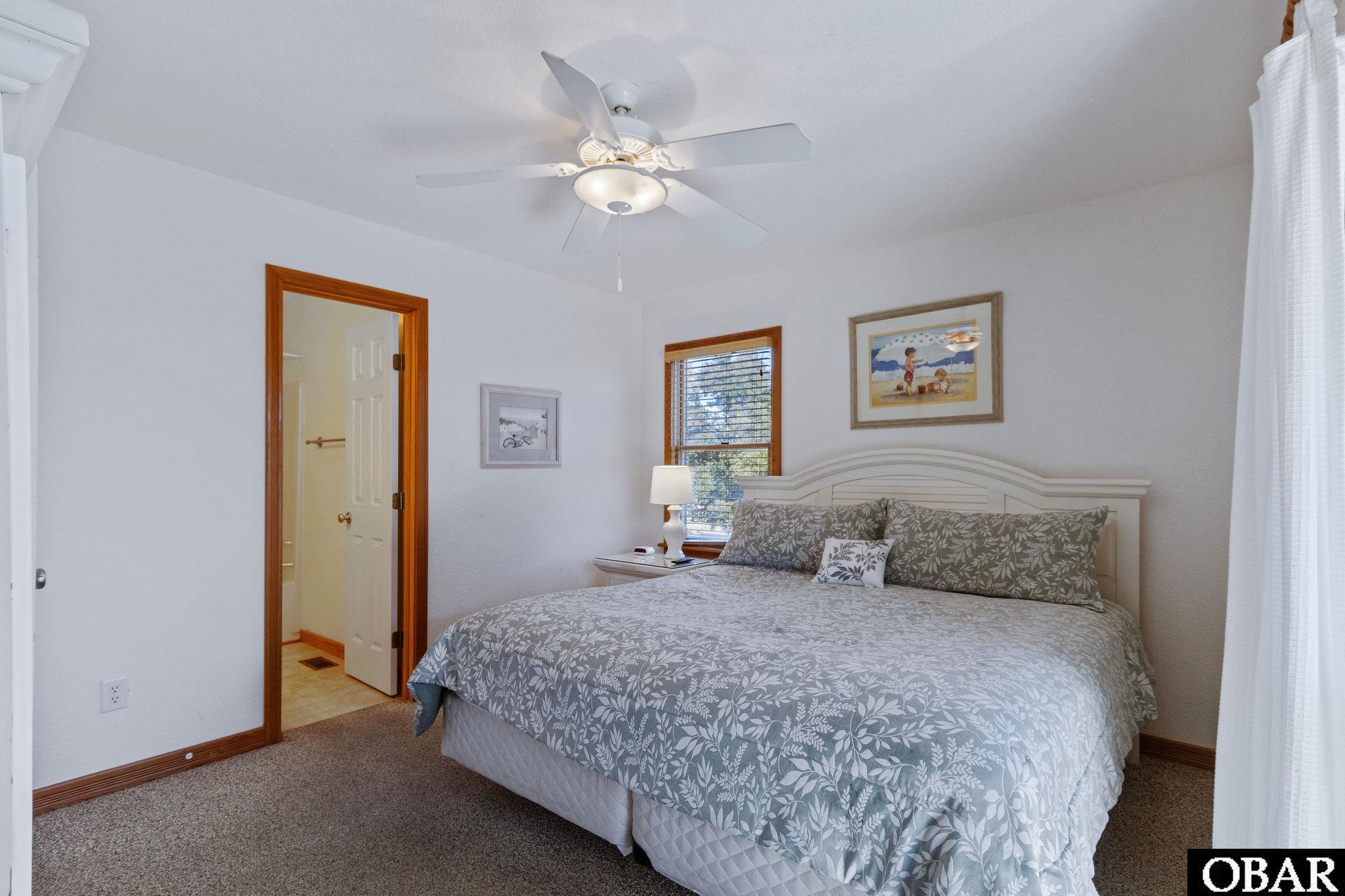 Bedroom with a large gray floral bed, white headboard, ceiling fan, and adjoining bathroom doorway.