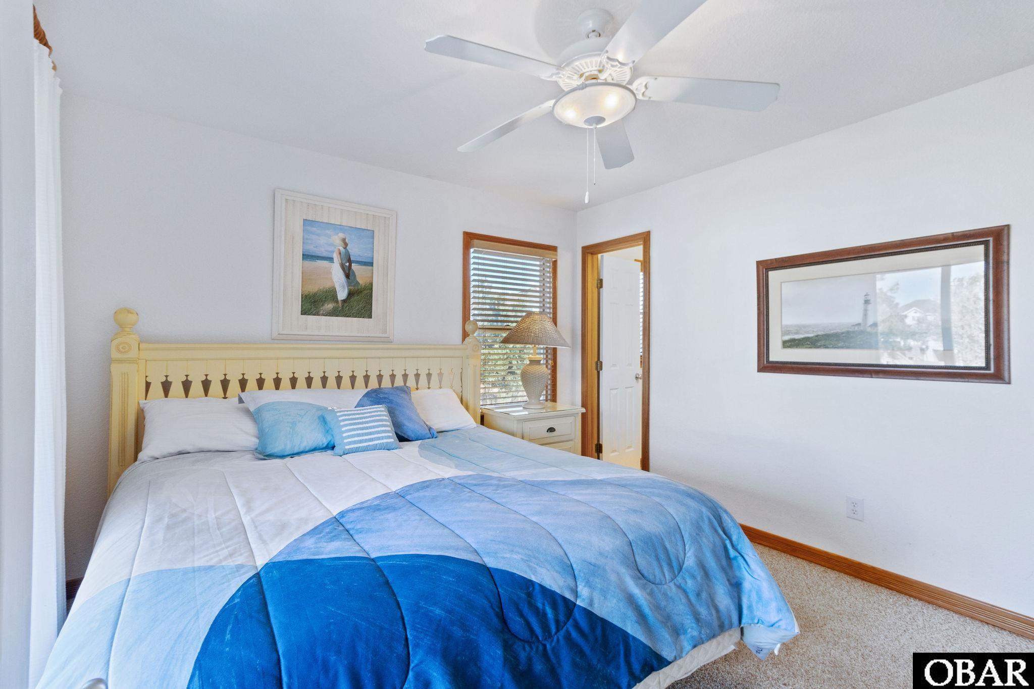 Bedroom with a cream spindle bed, blue-and-white quilt, coastal artwork, and wood-trimmed windows.
