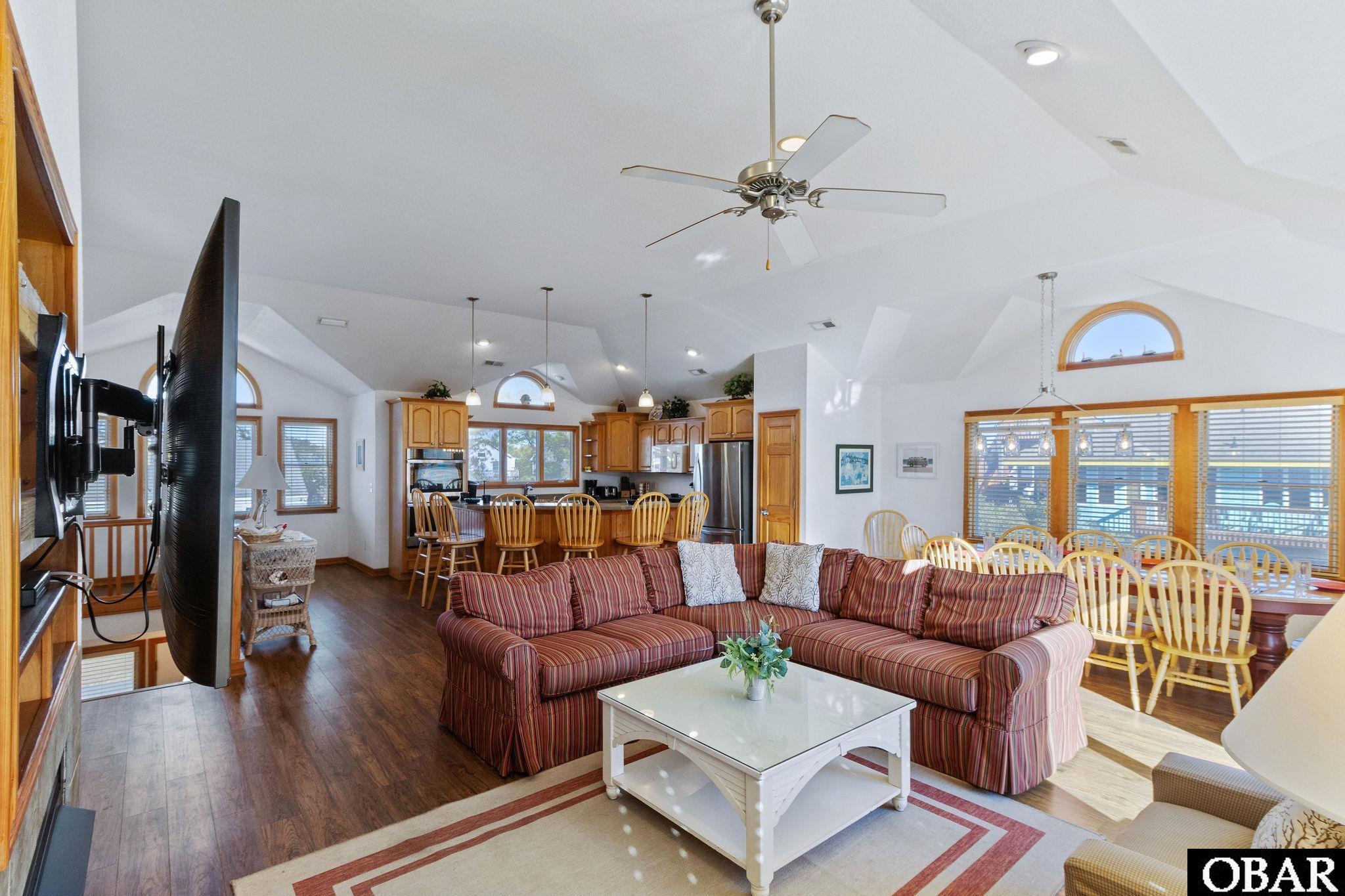 Open-plan living and dining area with a striped sectional, vaulted ceiling, and water visible through the windows.