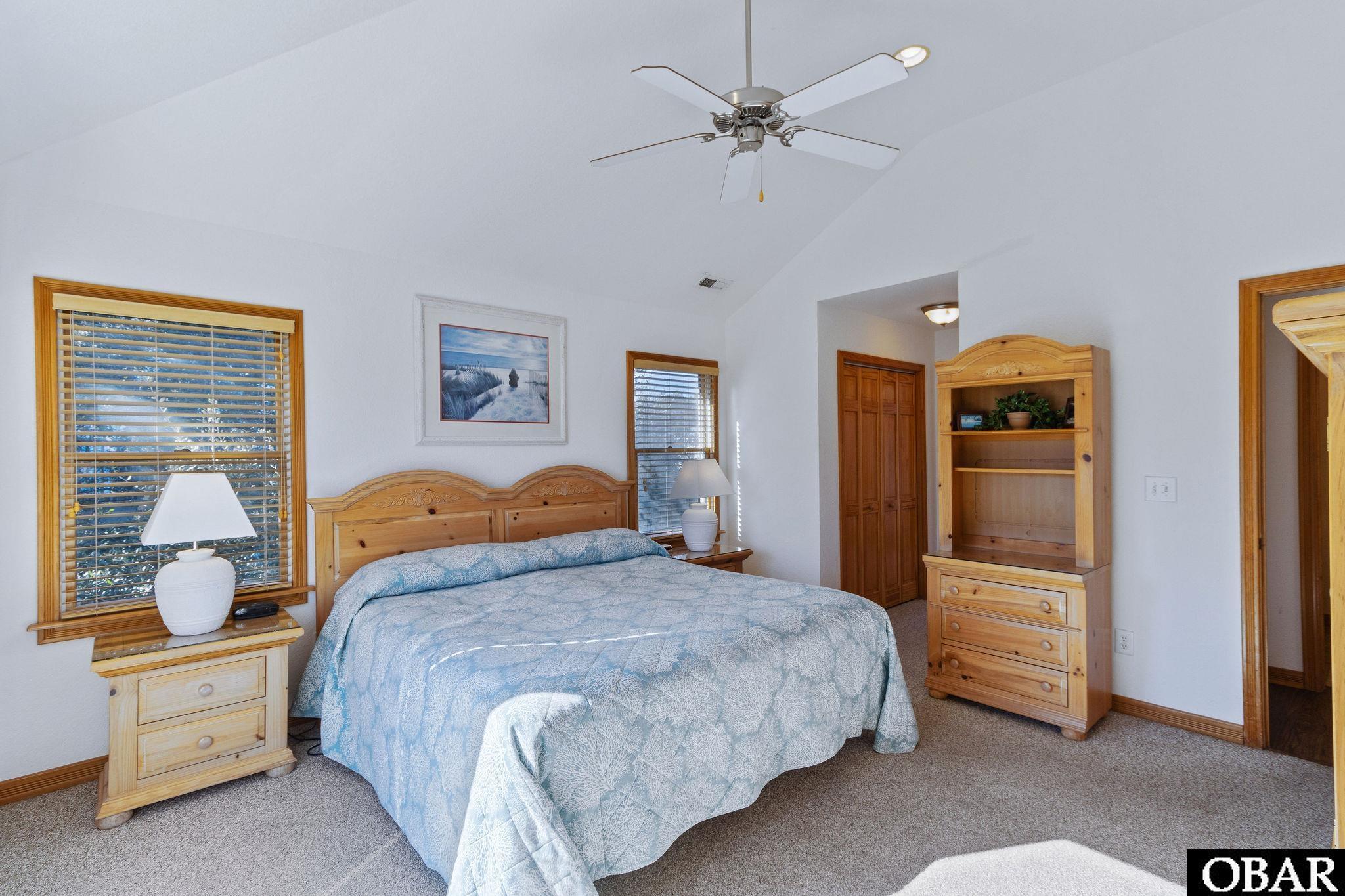 Furnished bedroom with a king bed, pale blue quilt, light wood furniture, and wood-trimmed windows.