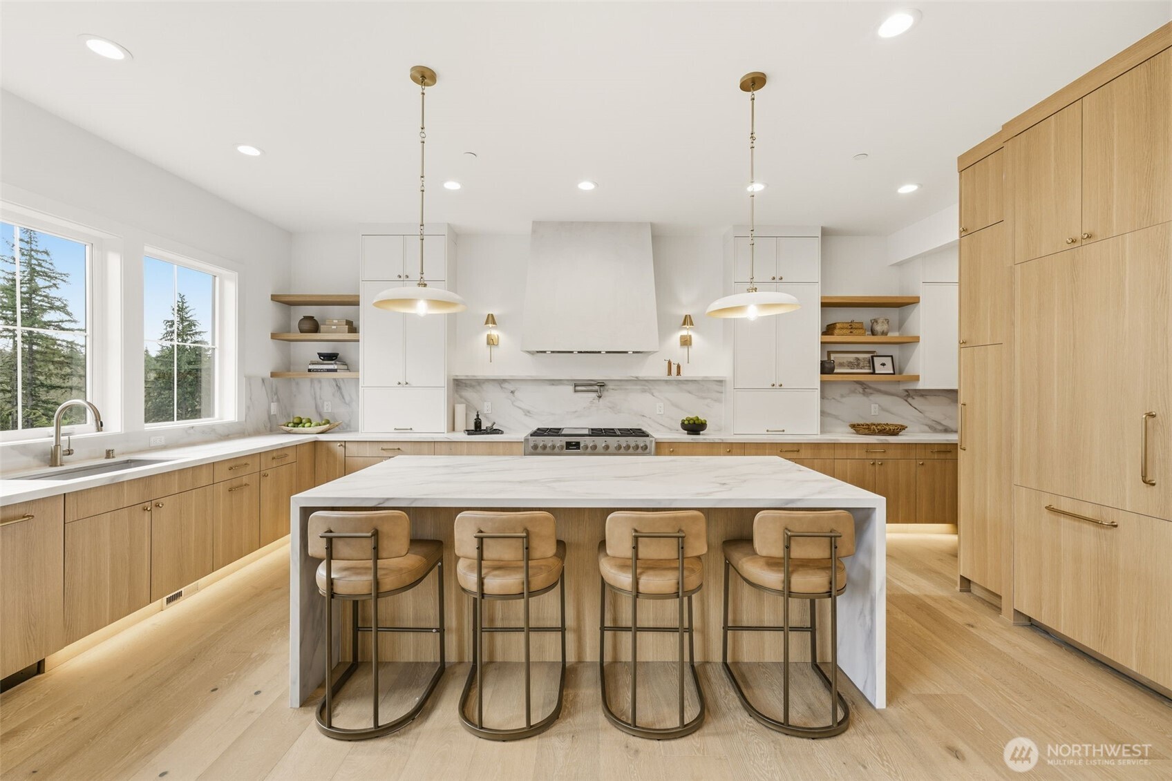 Kitchen with a marble waterfall island, four bar stools, light wood cabinetry, and windows overlooking evergreen trees.