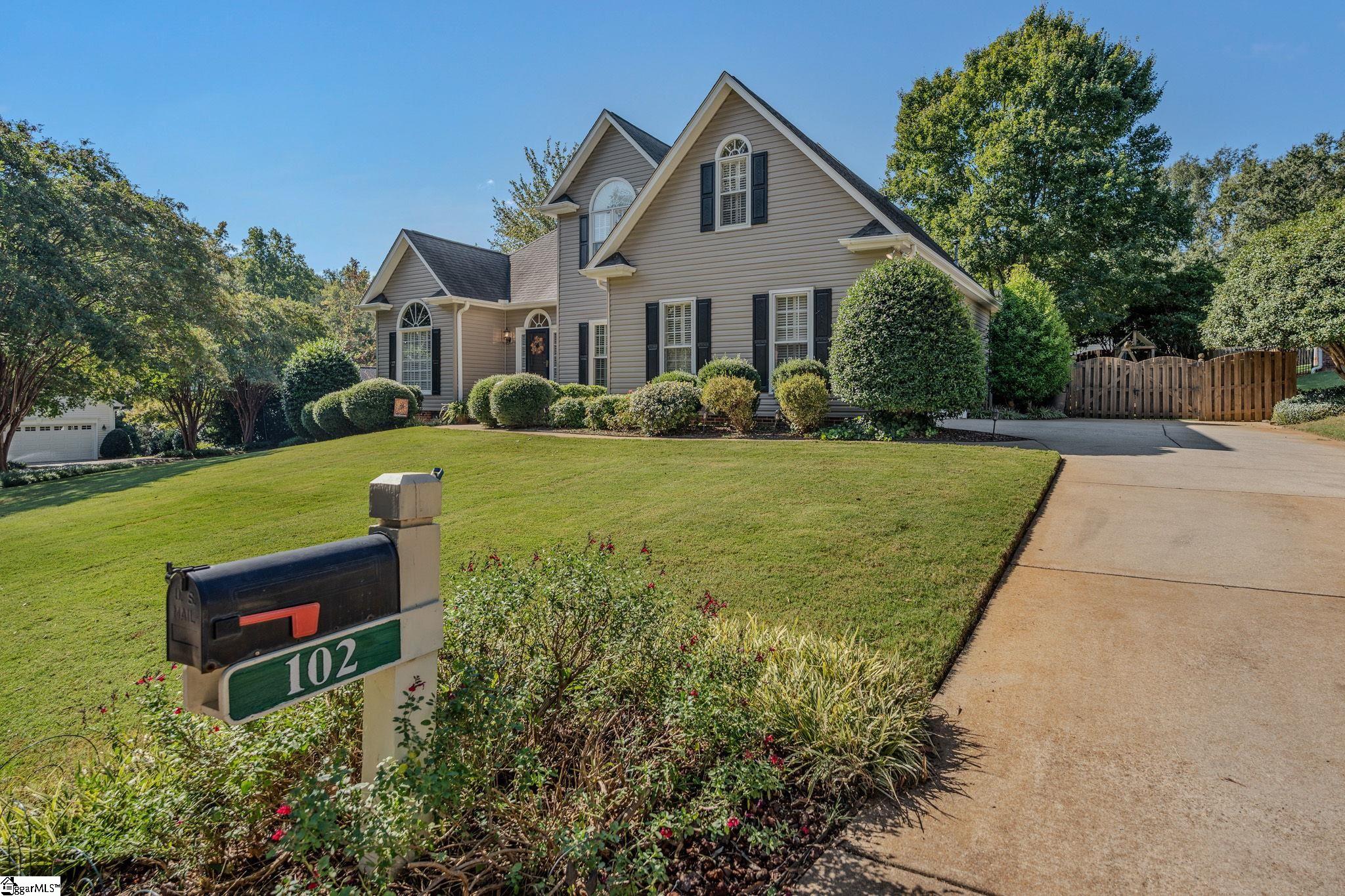 Two-story beige house with dark shutters, landscaped lawn, driveway, and mailbox marked 102.