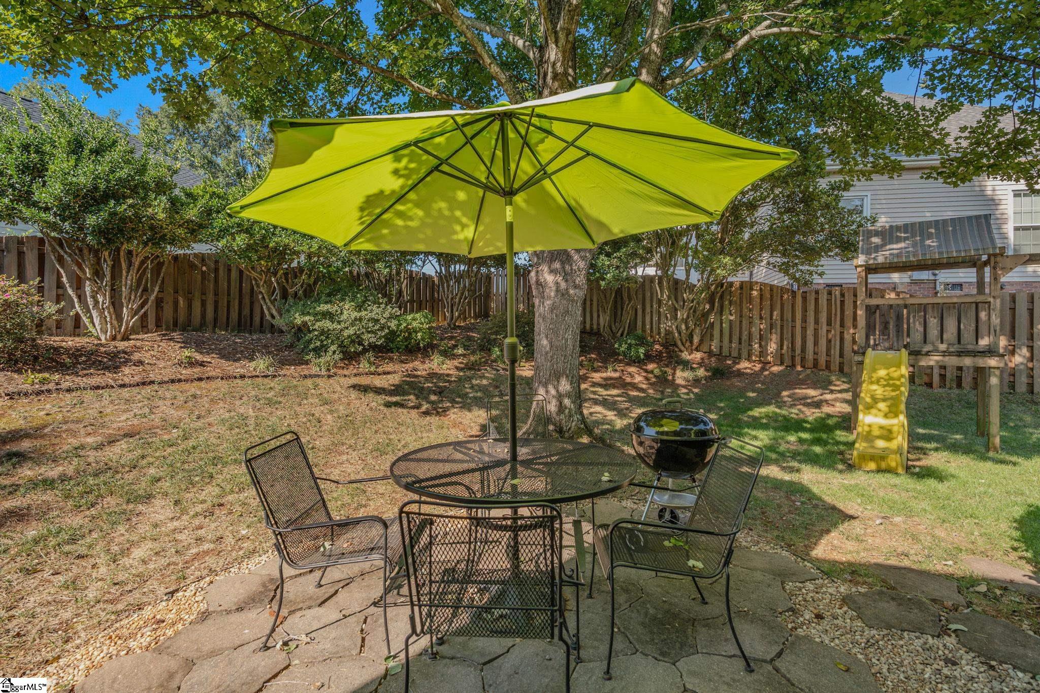 Backyard patio with metal seating beneath a bright green umbrella, beside a grill and children’s slide.