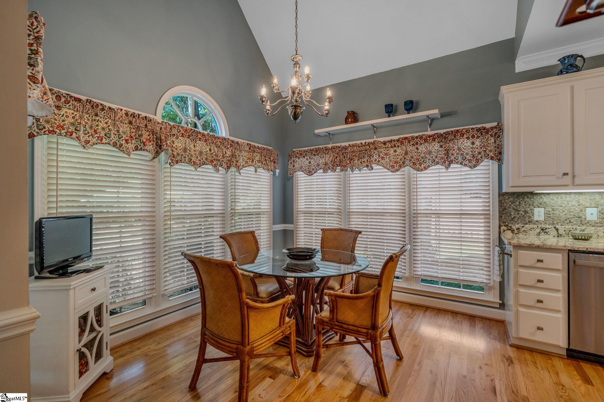 Dining nook with a round glass table, wicker chairs, chandelier, and large windows.