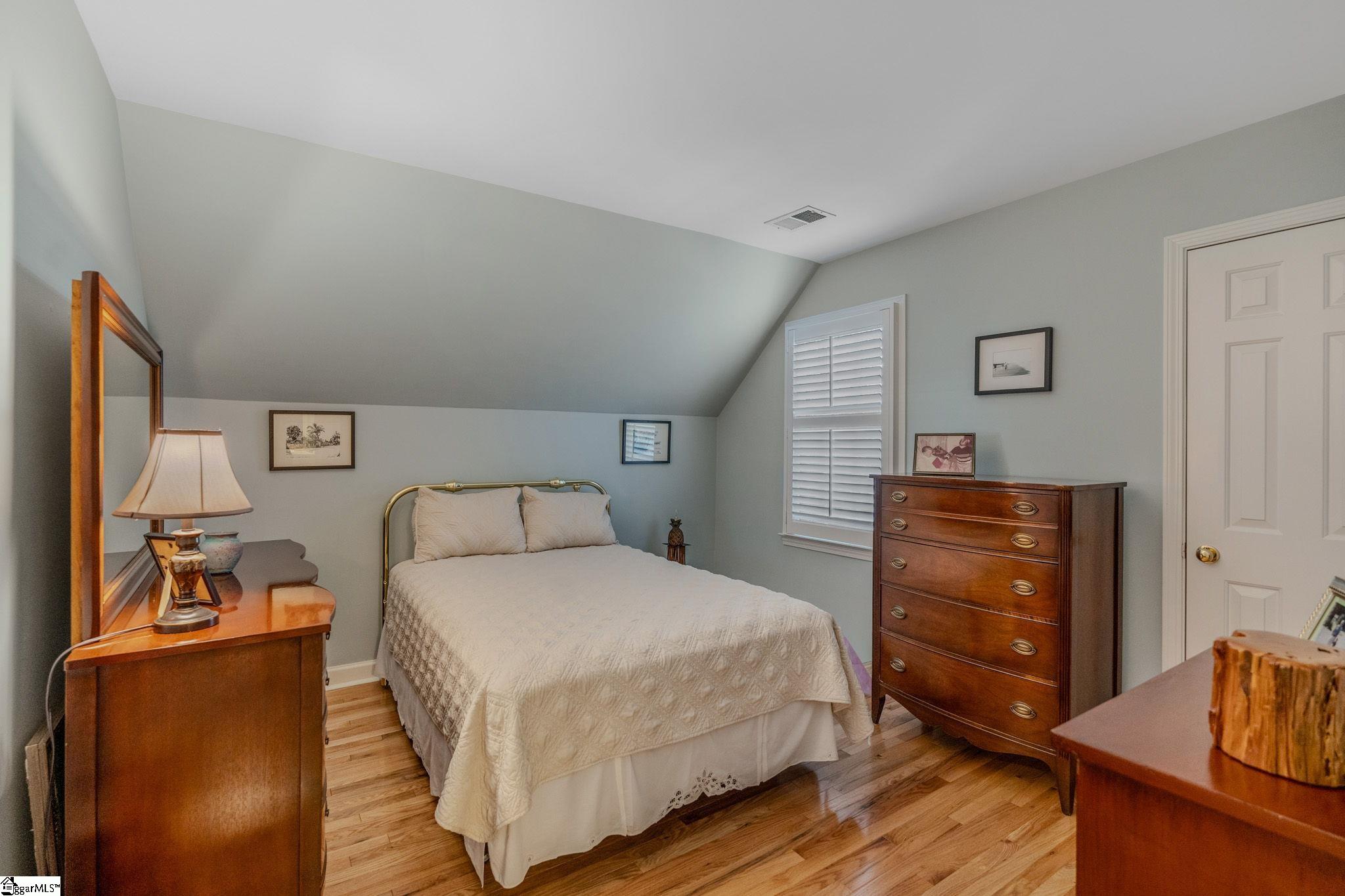 Furnished bedroom with a cream-covered bed, sloped ceiling, hardwood floors, and wooden dressers.