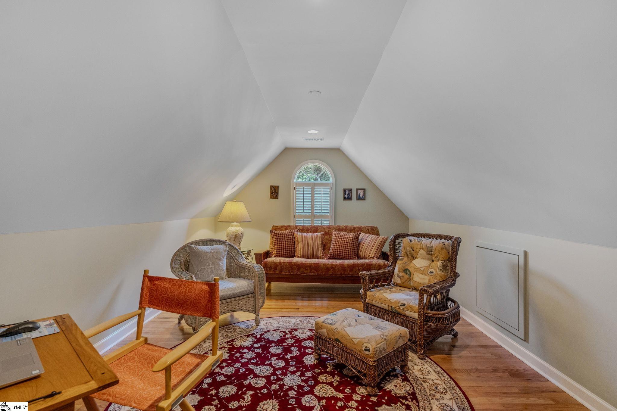 Attic sitting room with sloped ceilings, an arched shuttered window, wicker chairs, and a red floral rug.
