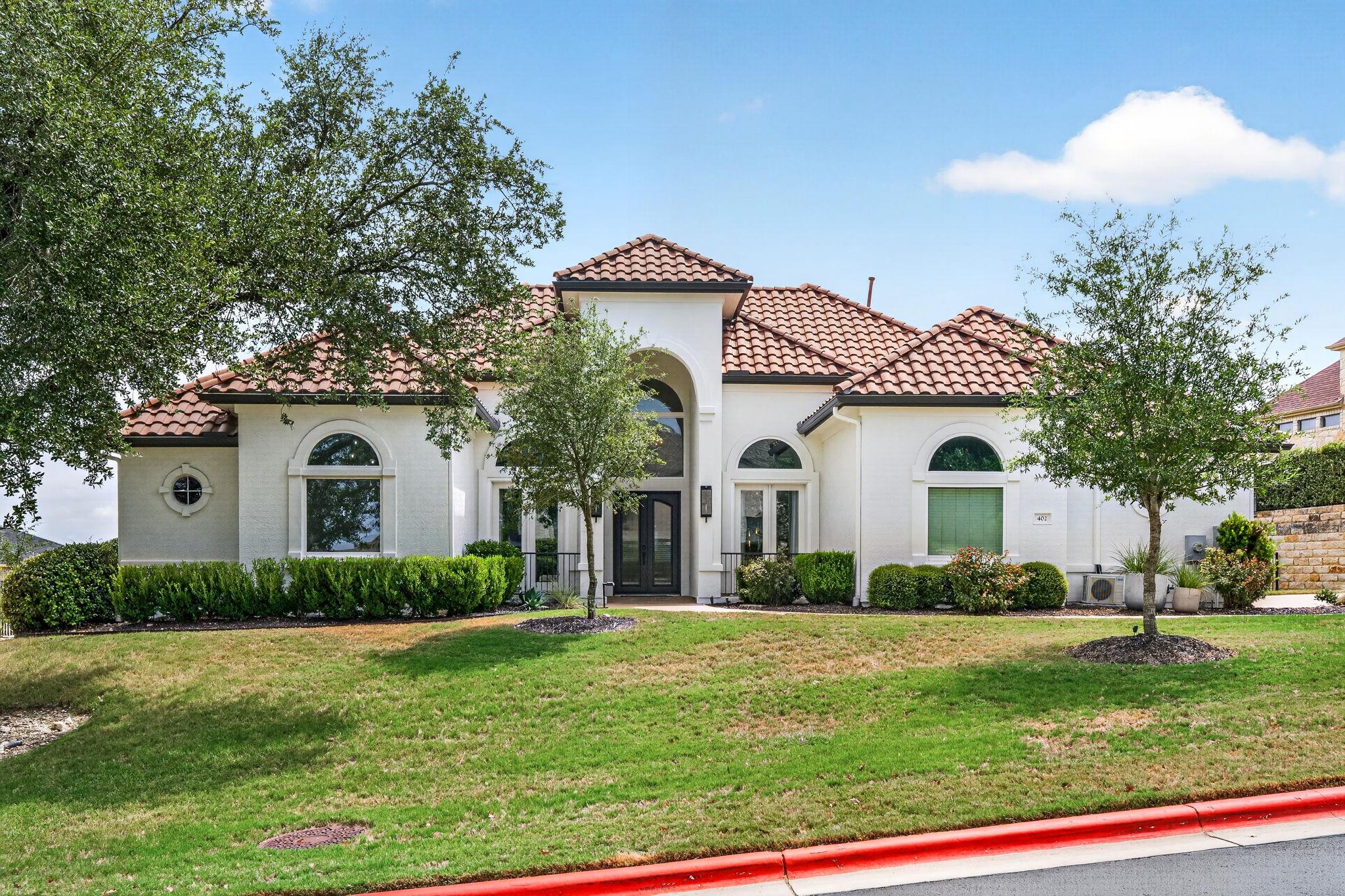 White stucco home with red tile roof, arched windows, mature trees, and a sloping lawn.