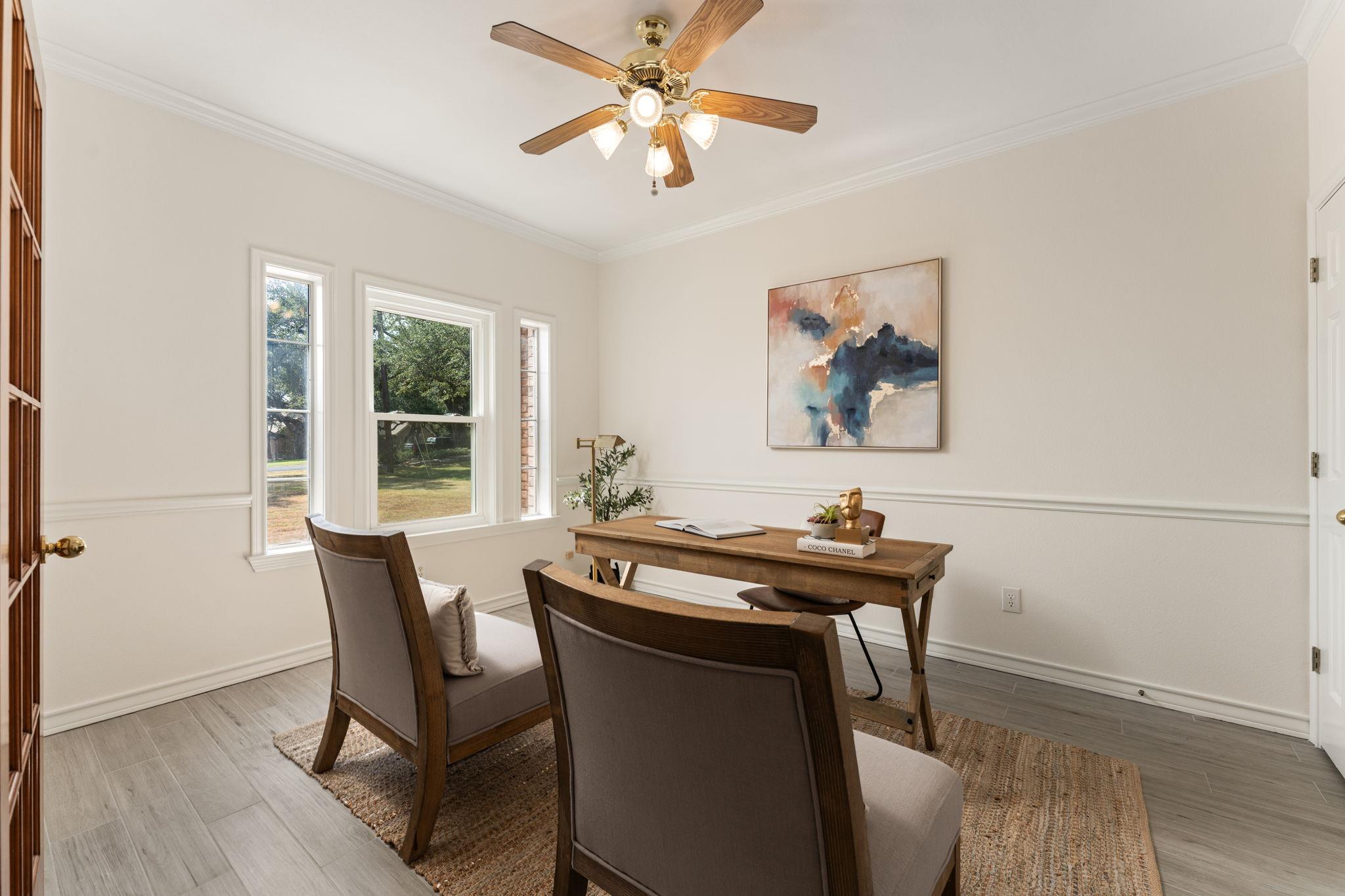 Home office with a wooden desk and chairs beside three windows overlooking a leafy yard.